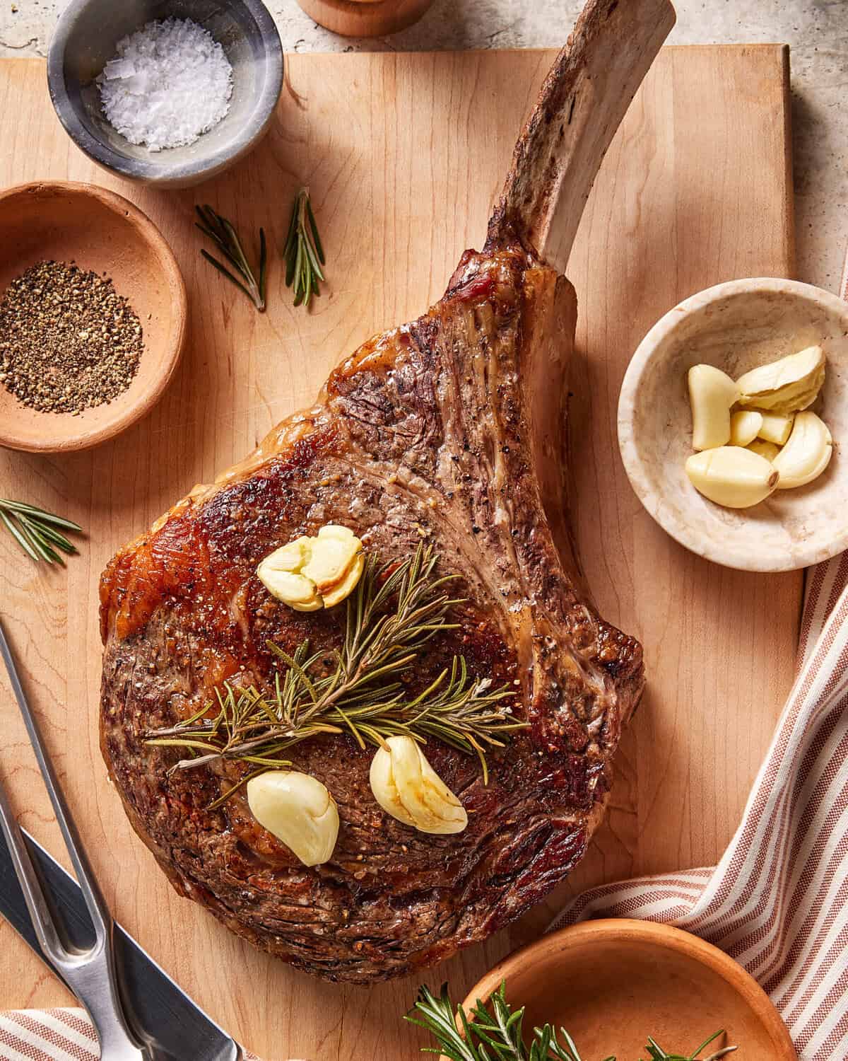 A cooked bone-in ribeye steak on a wooden cutting board, topped with rosemary and garlic cloves. Surrounding the steak are bowls of salt, pepper, more garlic, and sprigs of rosemary, with utensils and a striped napkin.