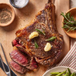 A cooked tomahawk steak with garlic cloves and rosemary on top, partially sliced, on a wooden cutting board. Surrounding are bowls of salt, pepper, fresh rosemary, and a plate of green beans.