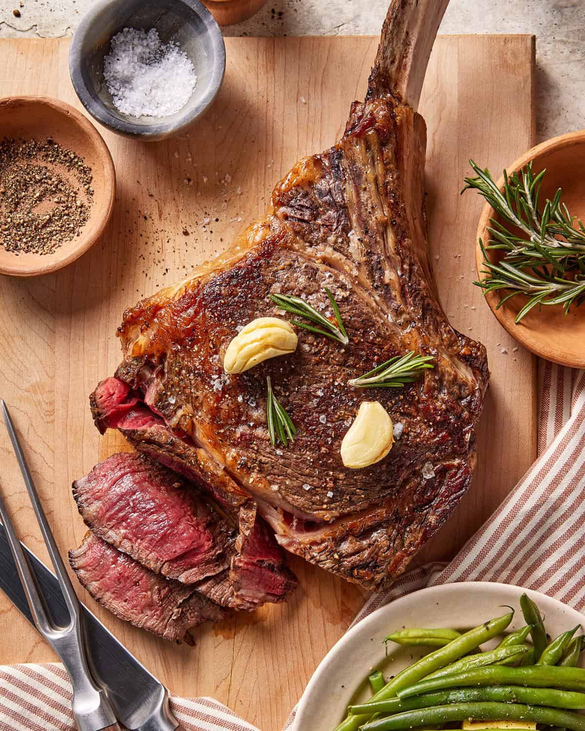 A cooked tomahawk steak with garlic cloves and rosemary on top, partially sliced, on a wooden cutting board. Surrounding are bowls of salt, pepper, fresh rosemary, and a plate of green beans.