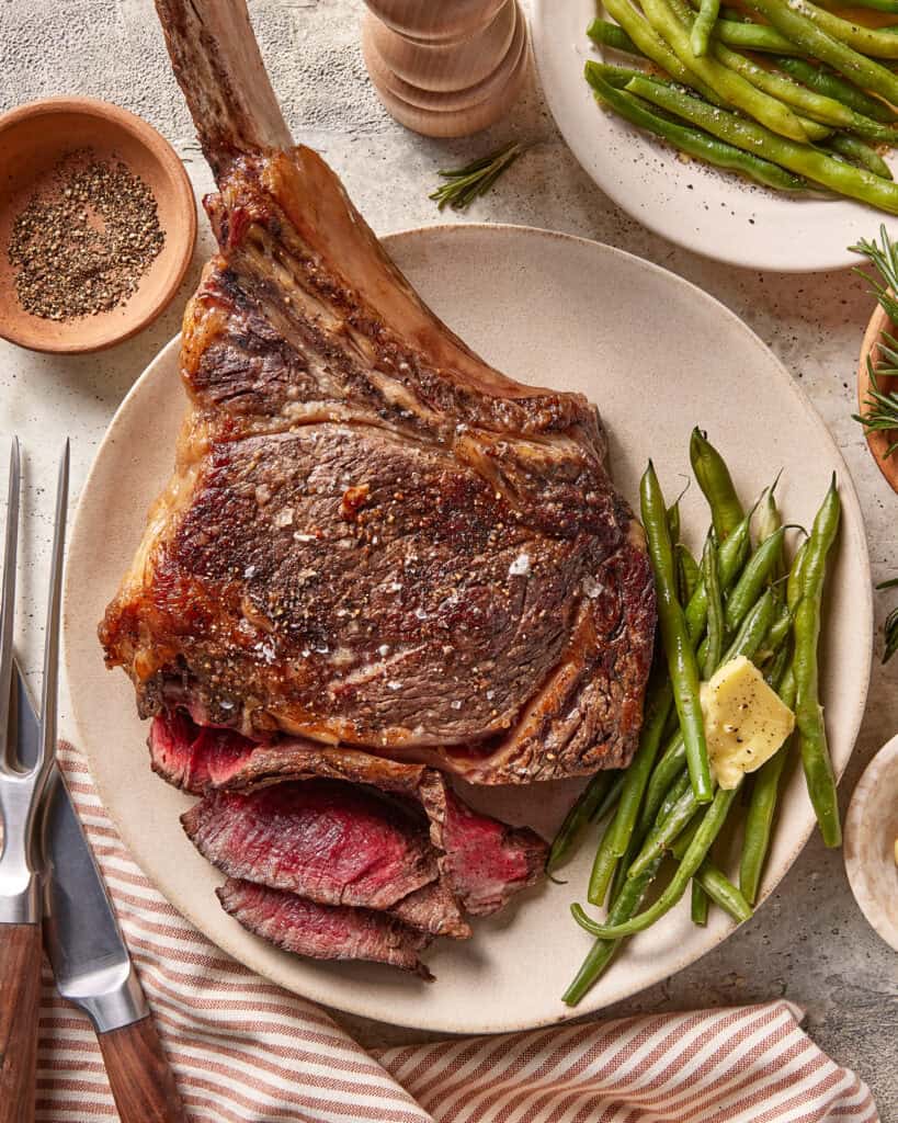 A plate with a large, bone-in steak, several slices cut, served with green beans and a pat of butter. Nearby are a fork, knife, pepper, salt, and a plate of more green beans. A striped napkin is underneath the plate.