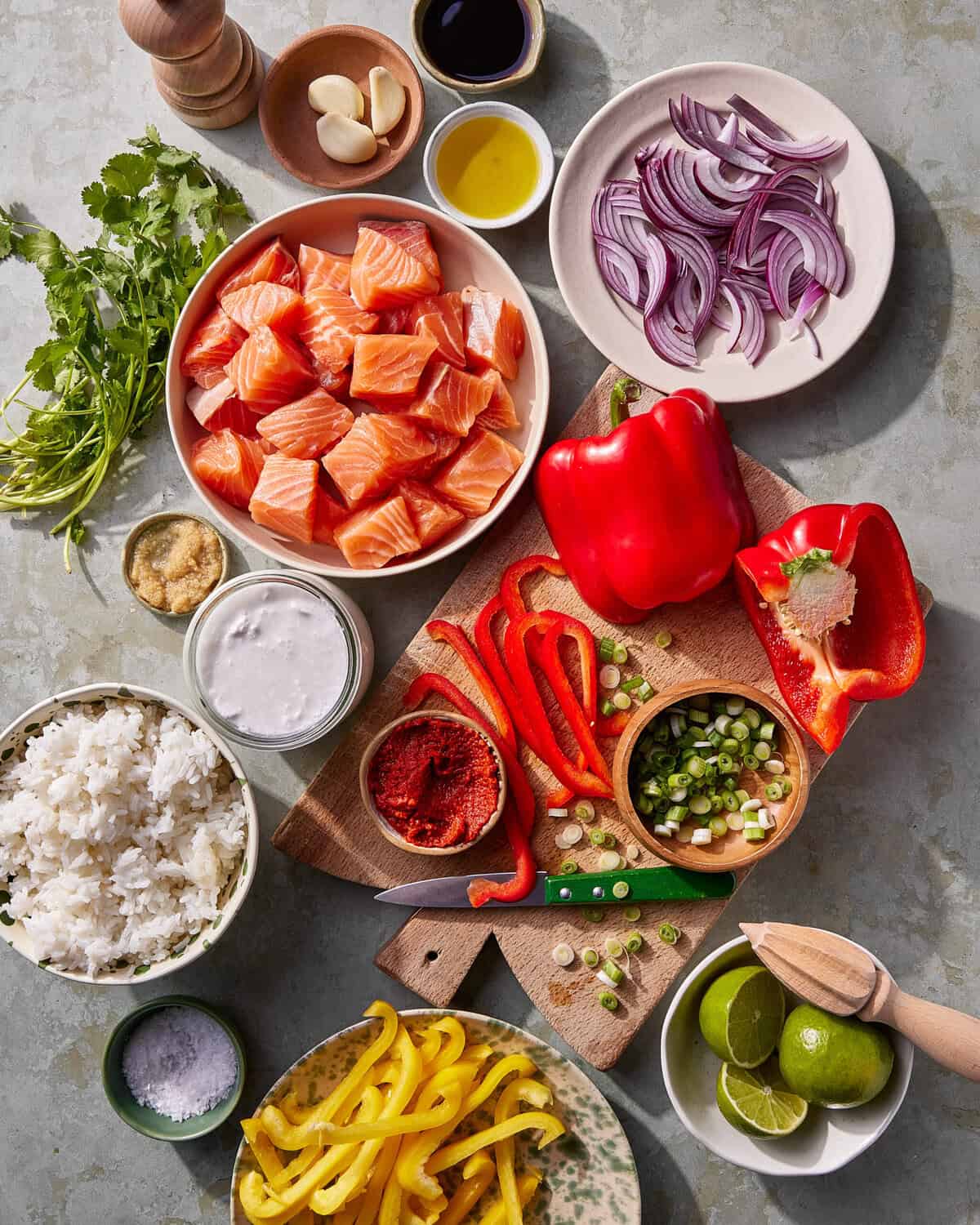 Ingredients for a meal are arranged on a table, including cubed raw salmon, sliced red and yellow bell peppers, chopped spring onions, red onion, cooked rice, limes, cilantro, garlic, seasonings, and small bowls of sauces.