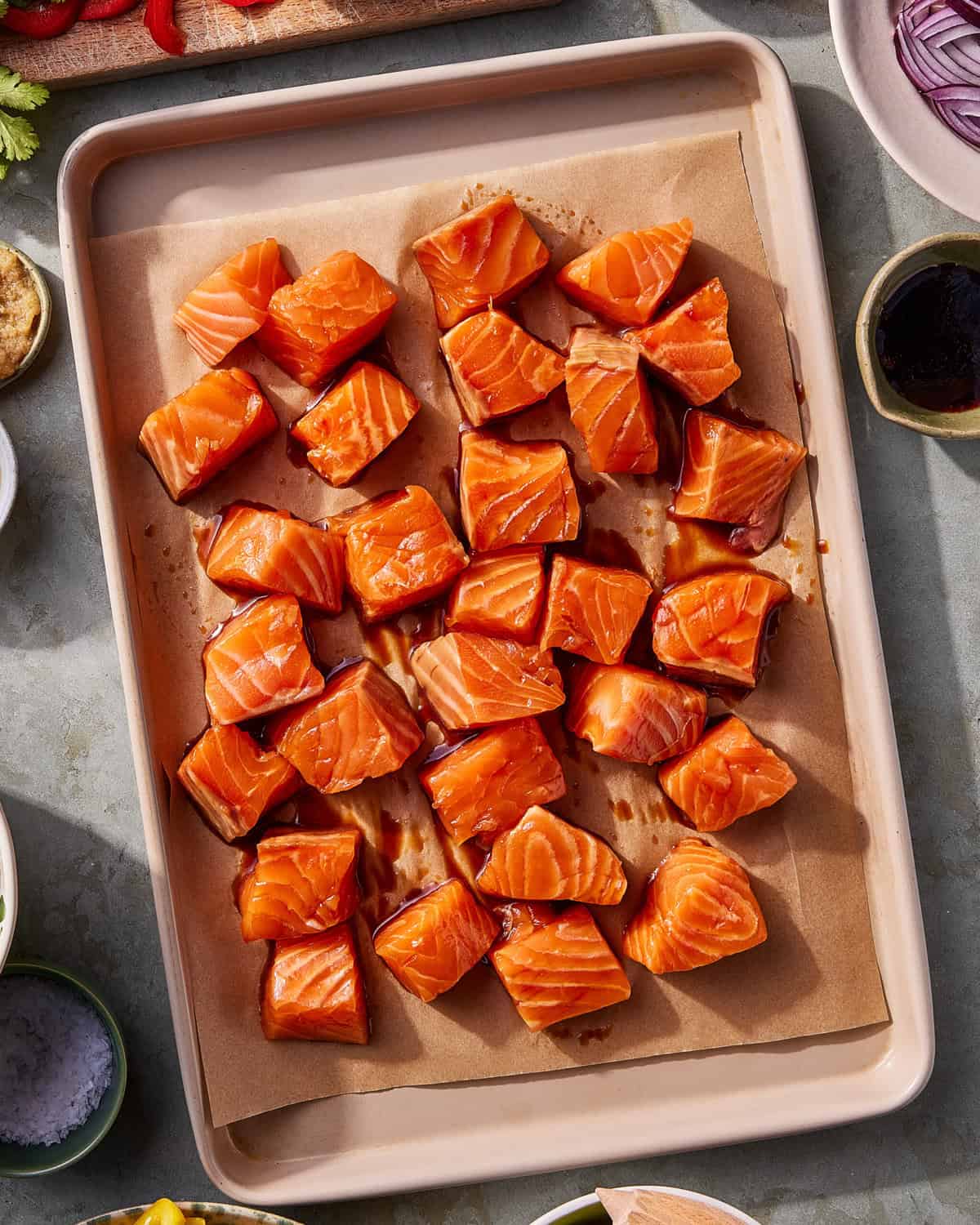 Cubes of raw salmon are arranged in rows on a parchment-lined baking sheet, surrounded by small bowls and ingredients on a gray countertop.