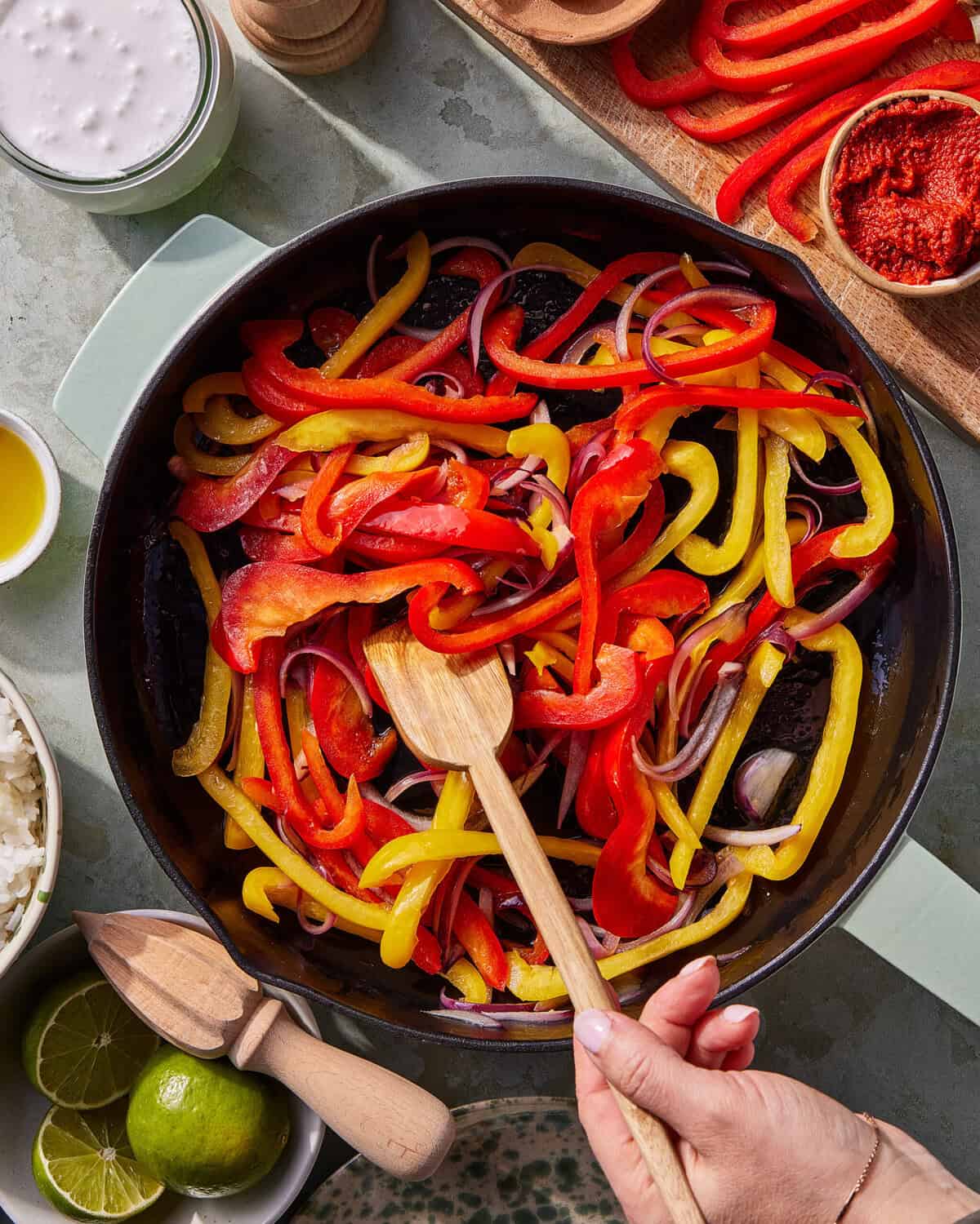 A hand stirs sliced red and yellow bell peppers and red onions in a skillet. Surrounding the skillet are bowls of rice, olive oil, coconut milk, tomato paste, and halved limes on a light countertop.