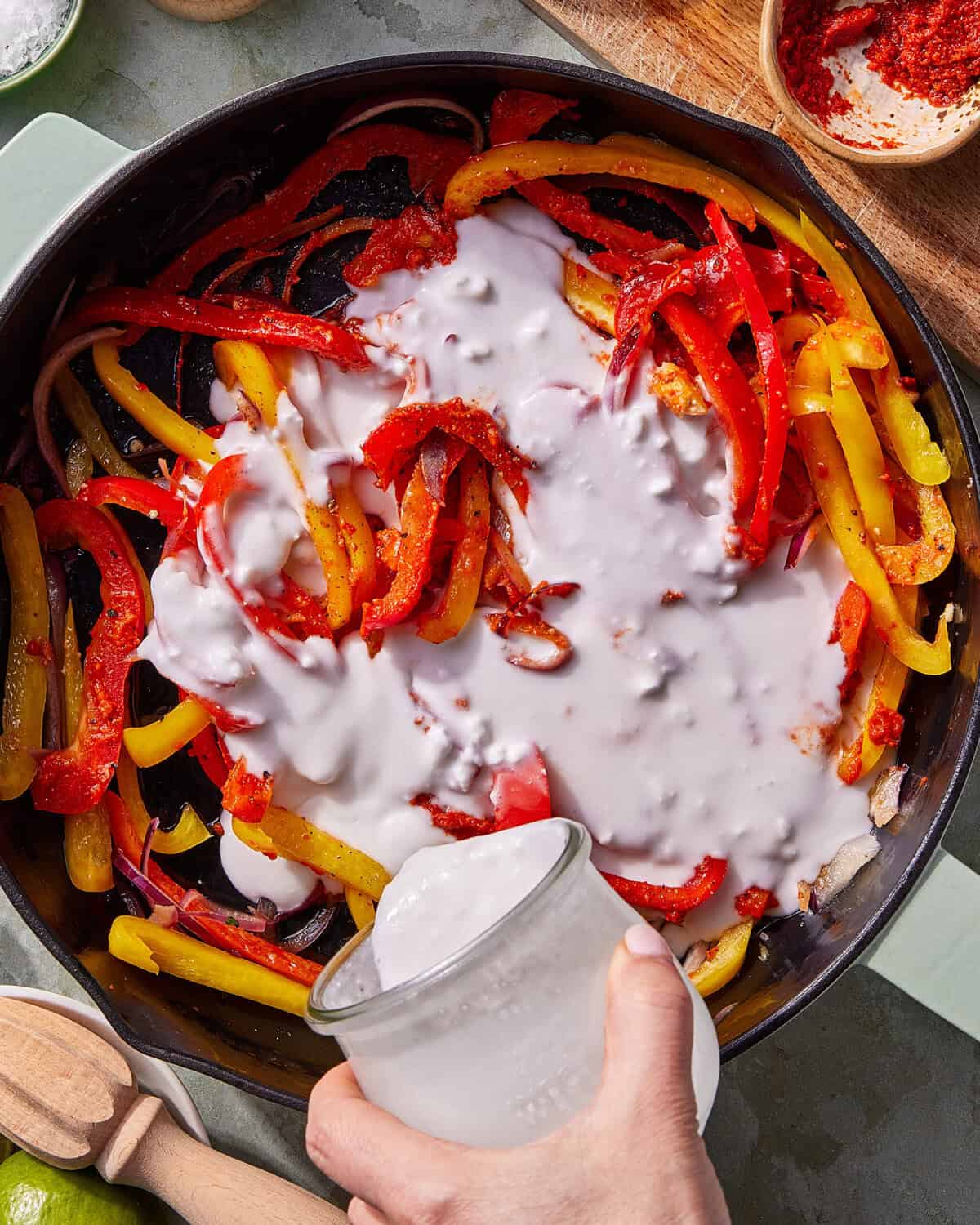 A hand pours coconut milk from a jar into a skillet filled with sautéed red and yellow bell peppers and onions, with seasonings visible. A wooden citrus reamer and lime are nearby.