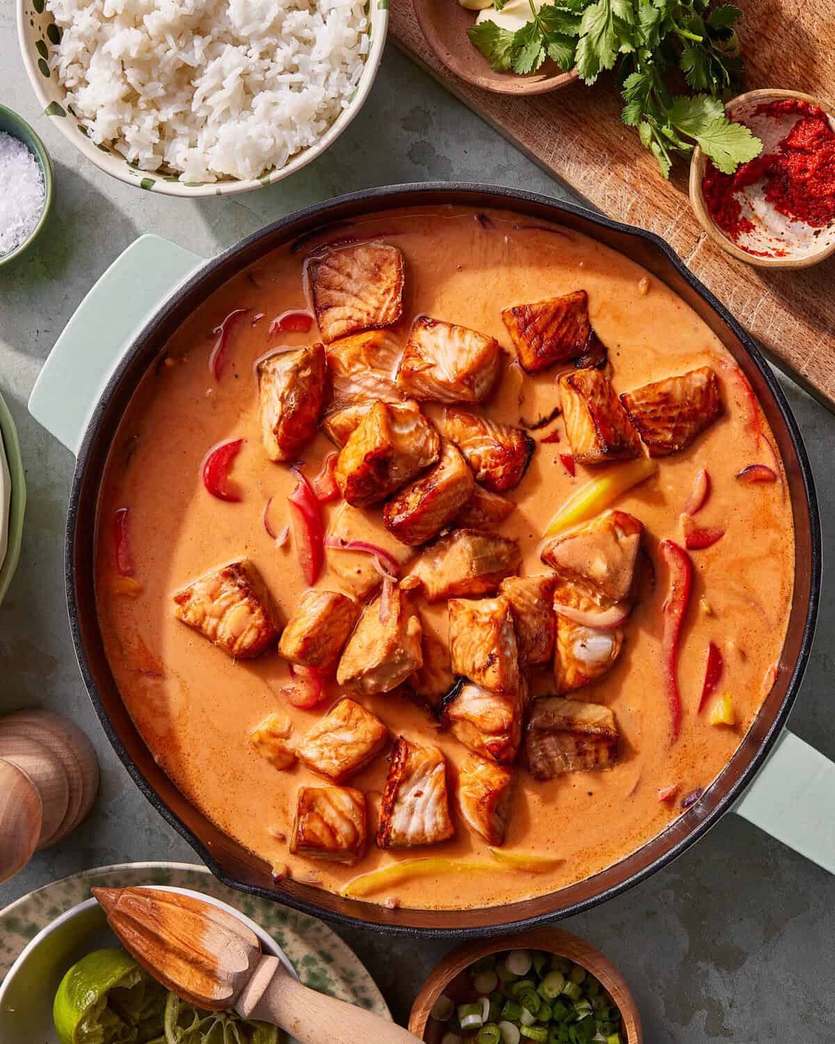 A pot of creamy, orange Thai curry with chunks of cooked salmon and red bell pepper slices, surrounded by bowls of rice, herbs, lime wedges, scallions, and spices on a gray countertop.