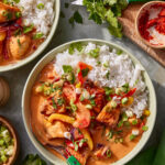 A bowl of white rice topped with a creamy curry containing pieces of fish, bell peppers, and herbs, garnished with chopped green onions and cilantro. Fresh ingredients and a bowl of spices are nearby on the table.