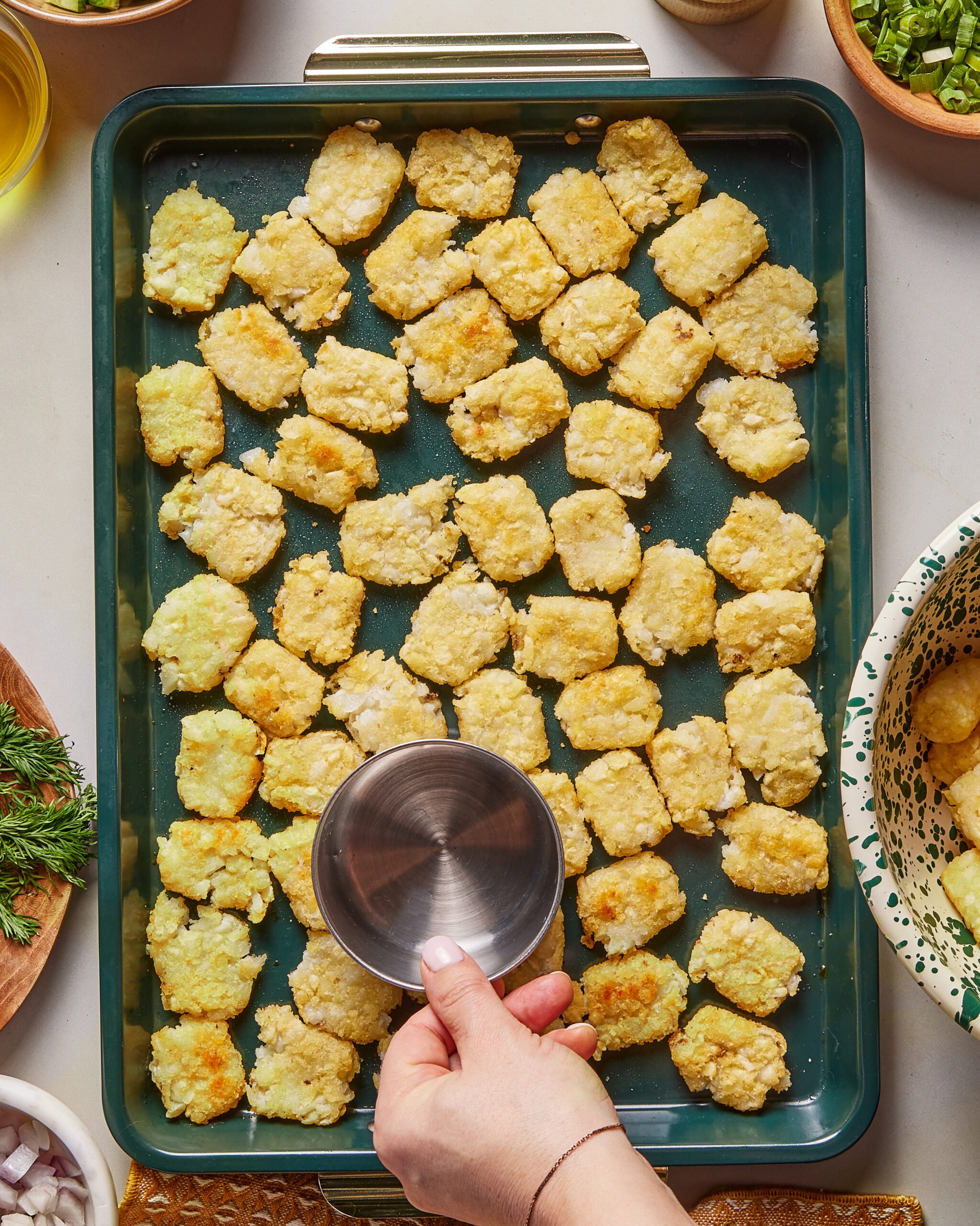 A hand is holding a metal measuring cup over a baking sheet filled with evenly arranged tater tots, surrounded by bowls of herbs and chopped vegetables.