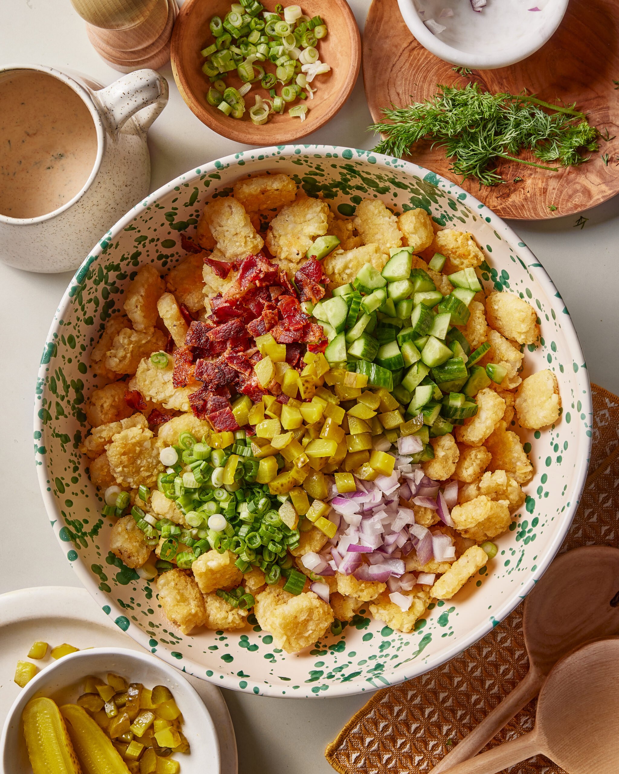 A large bowl filled with tater tots, topped with chopped bacon, cucumbers, pickles, red onion, and green onions. Around the bowl are fresh dill, sliced green onions, sauce, pepper, and extra pickles.