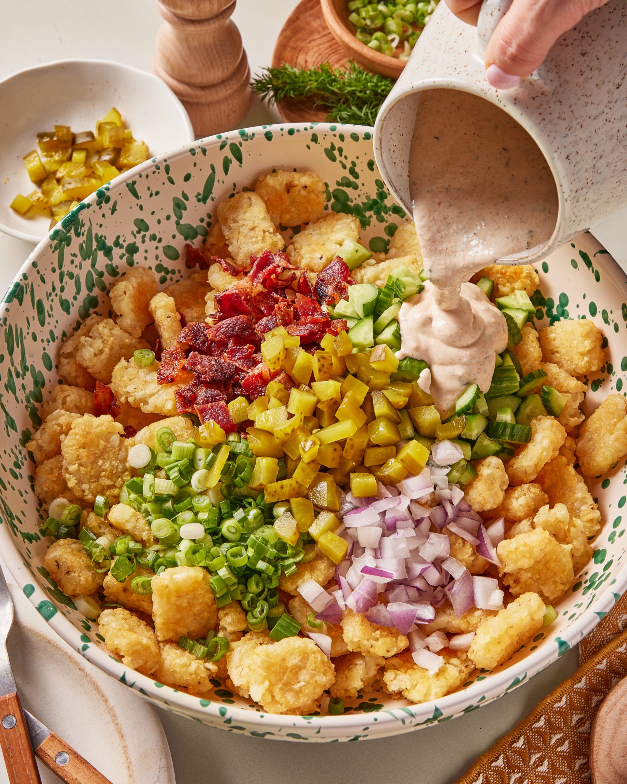 A large bowl of tater tots topped with chopped bacon, pickles, cucumber, green onions, and red onion, with dressing being poured over the ingredients. Sliced pickles and seasonings are nearby.