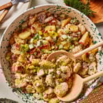 A bowl of creamy potato salad with chopped green onions, diced pickles, fresh dill, and crispy bacon bits, served with wooden salad utensils on a patterned plate. Forks, a glass of water, and herbs are nearby.