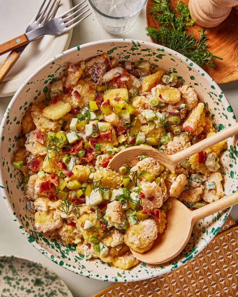 A bowl of creamy potato salad with chopped green onions, diced pickles, fresh dill, and crispy bacon bits, served with wooden salad utensils on a patterned plate. Forks, a glass of water, and herbs are nearby.