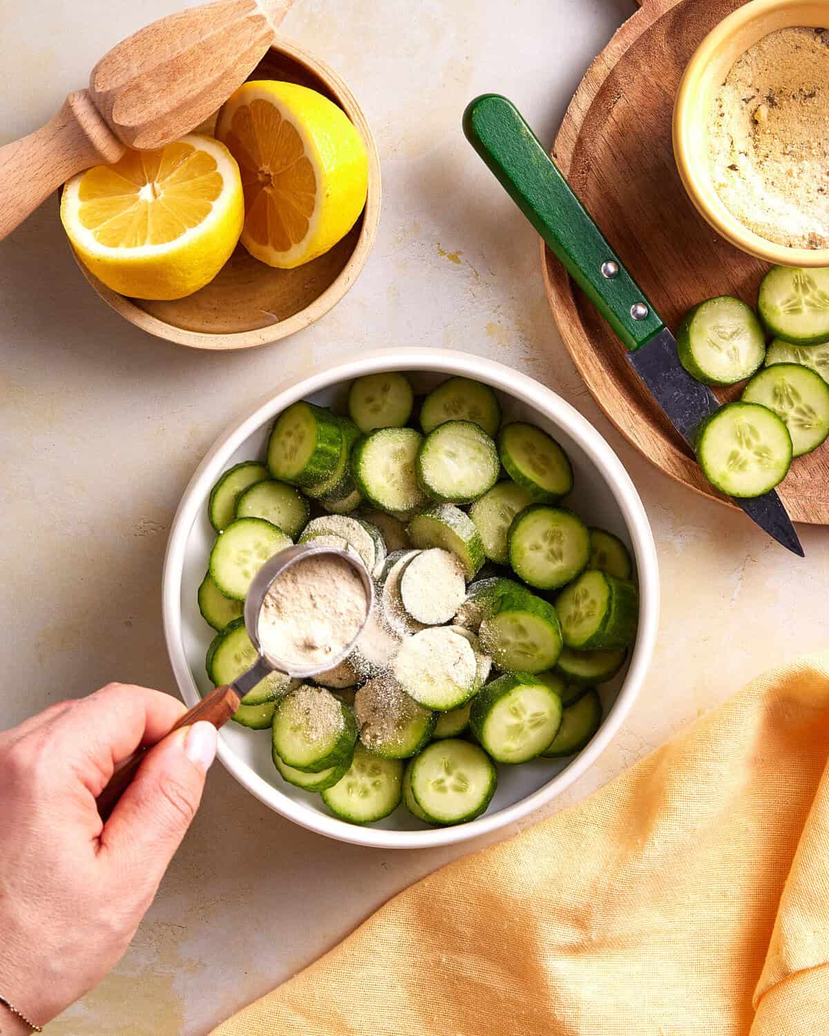 A hand sprinkles a powdery seasoning onto sliced cucumbers in a bowl. Nearby are lemon halves, a wooden citrus juicer, a knife, more cucumber slices, and a small bowl of additional seasoning. A yellow cloth is at the bottom.