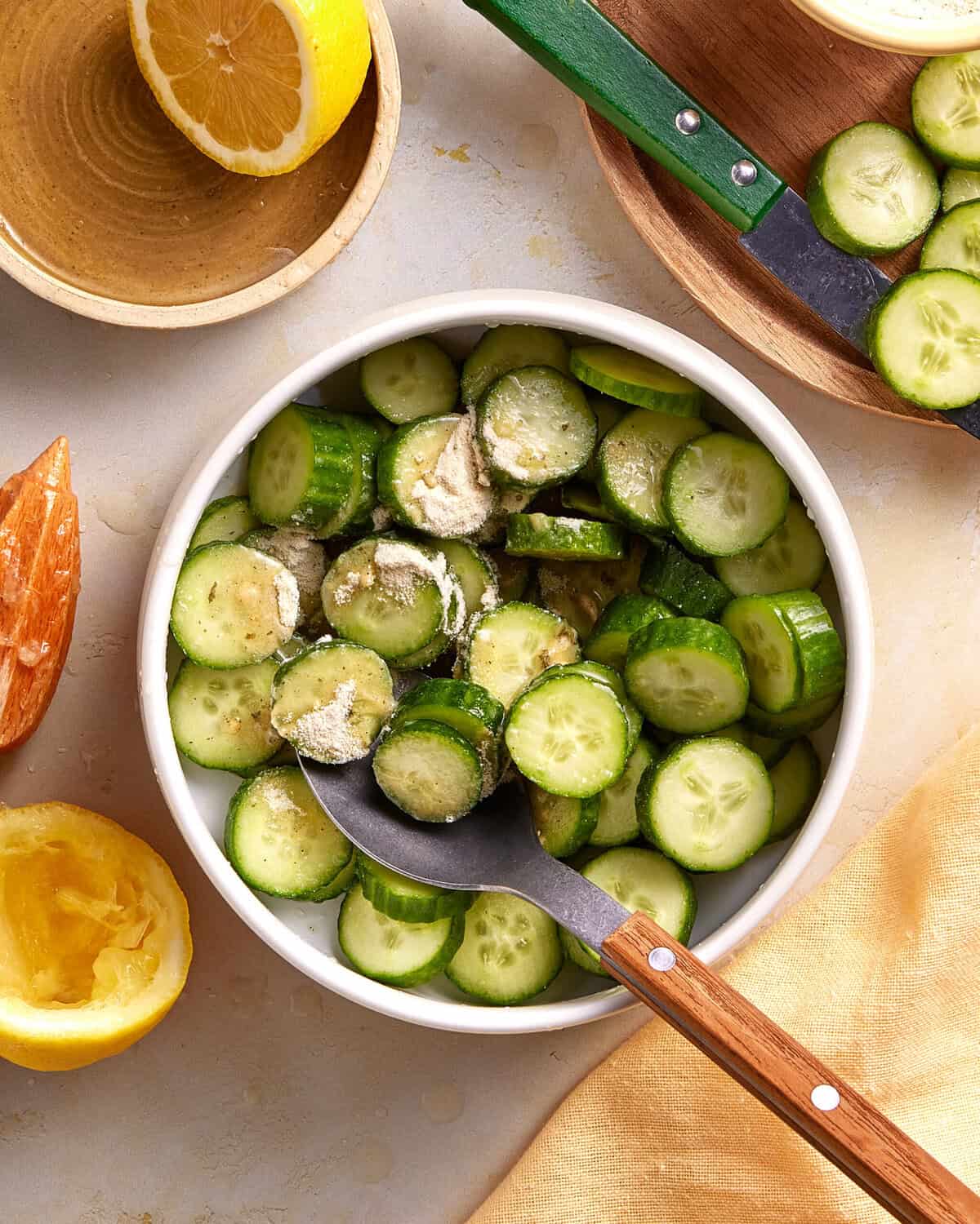 A white bowl filled with sliced cucumbers and a powder seasoning, with a spoon inside. Surrounding the bowl are halved lemons, a wooden bowl, and a wooden board with more sliced cucumbers.