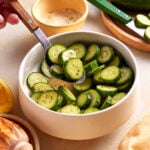 A hand holds a wooden spoon scooping sliced cucumbers from a white bowl. Other ingredients like a small bowl of seasoning, whole cucumbers, and a lemon are visible on the table.