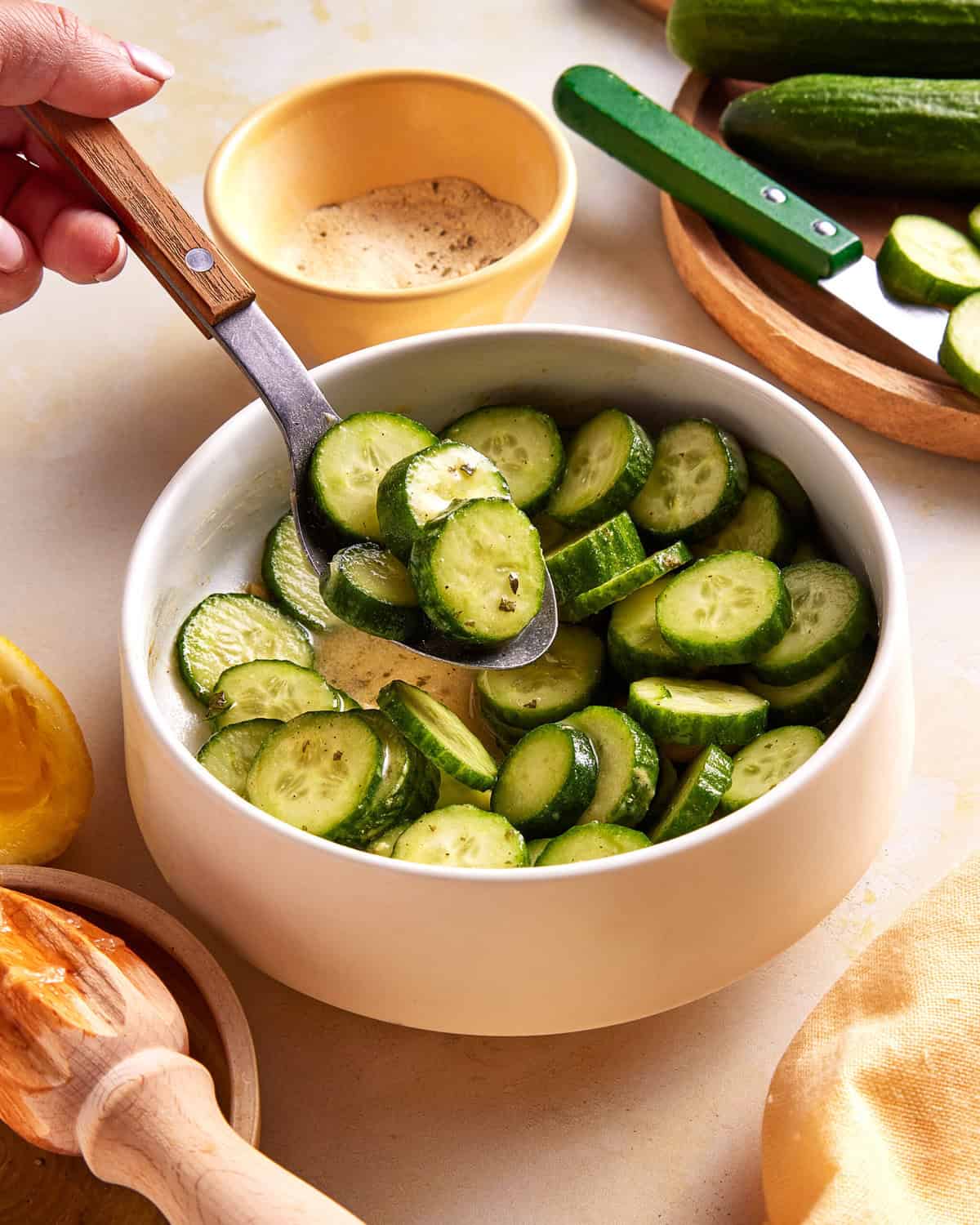 A hand holds a wooden spoon scooping sliced cucumbers from a white bowl. Other ingredients like a small bowl of seasoning, whole cucumbers, and a lemon are visible on the table.