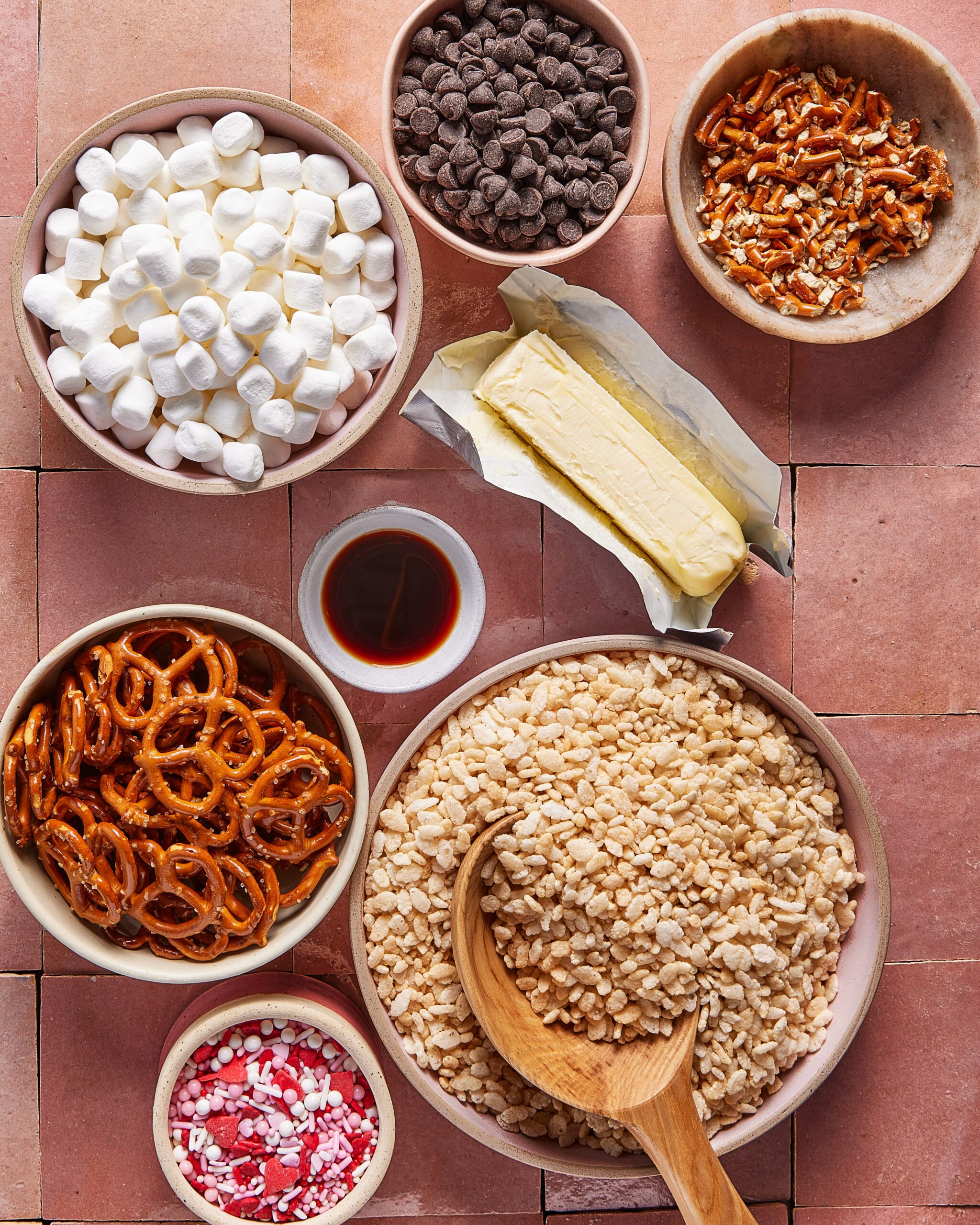Bowls filled with mini marshmallows, chocolate chips, chopped pretzels, pretzel twists, vanilla extract, peppermint bits, rice cereal, and a stick of butter on a pink tiled surface with a wooden spoon.