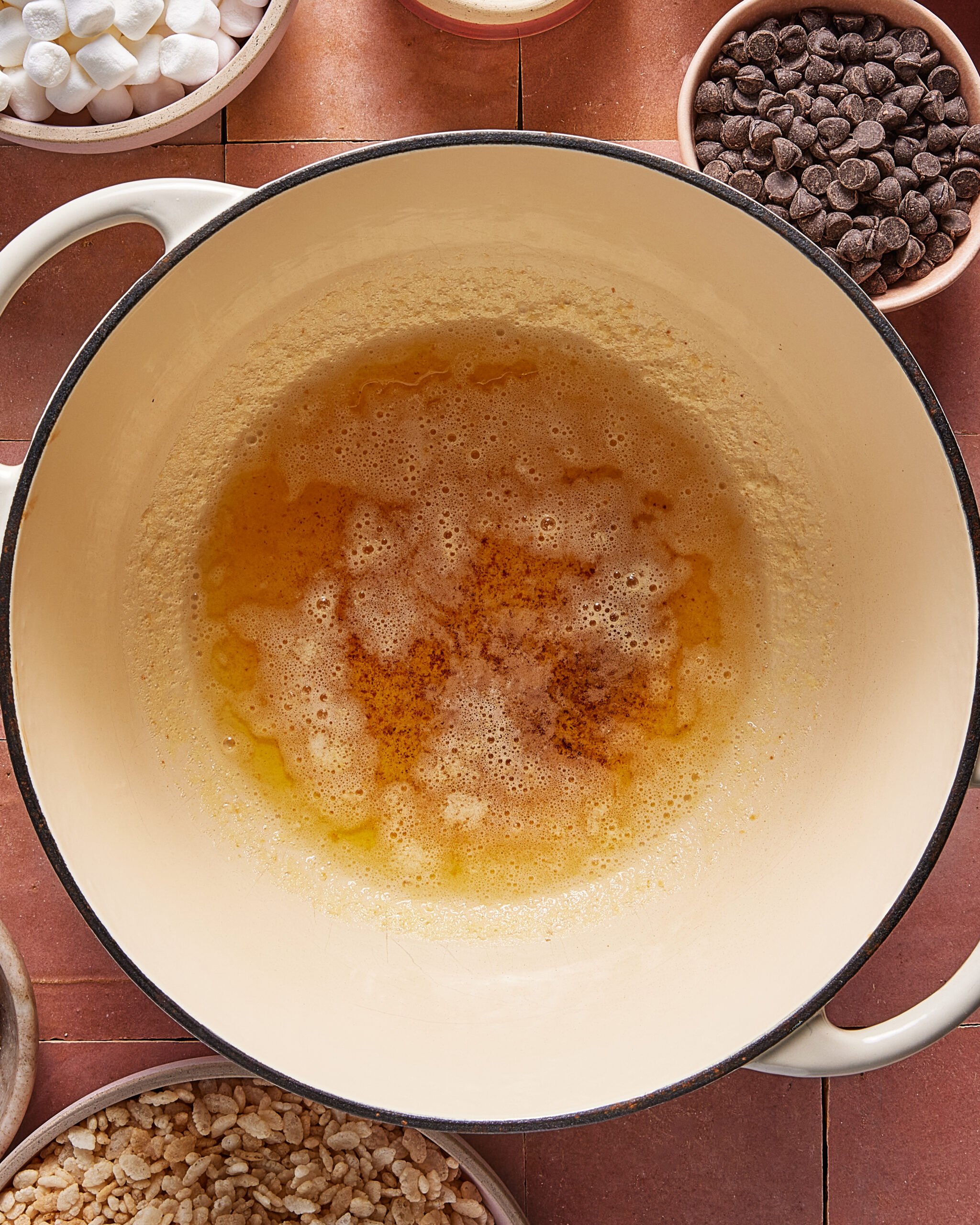 A large pot with melted and browned butter sits in the center, surrounded by bowls of chocolate chips, mini marshmallows, and puffed rice cereal on a reddish-brown tile surface.
