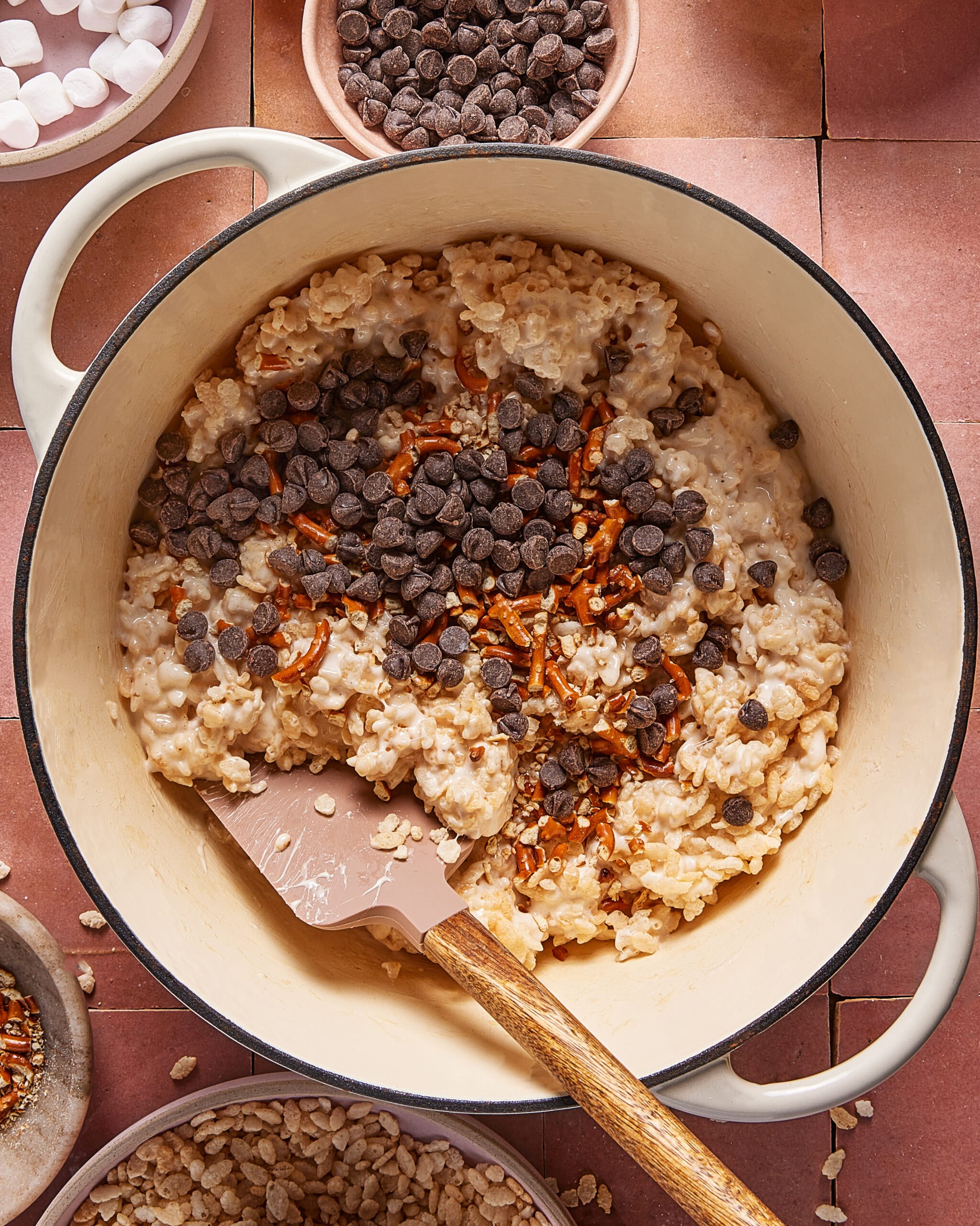 A pot filled with a mixture of melted marshmallows and rice cereal, topped with chocolate chips and pretzel pieces. A spatula with a wooden handle rests inside the pot. Small bowls of ingredients surround the pot.