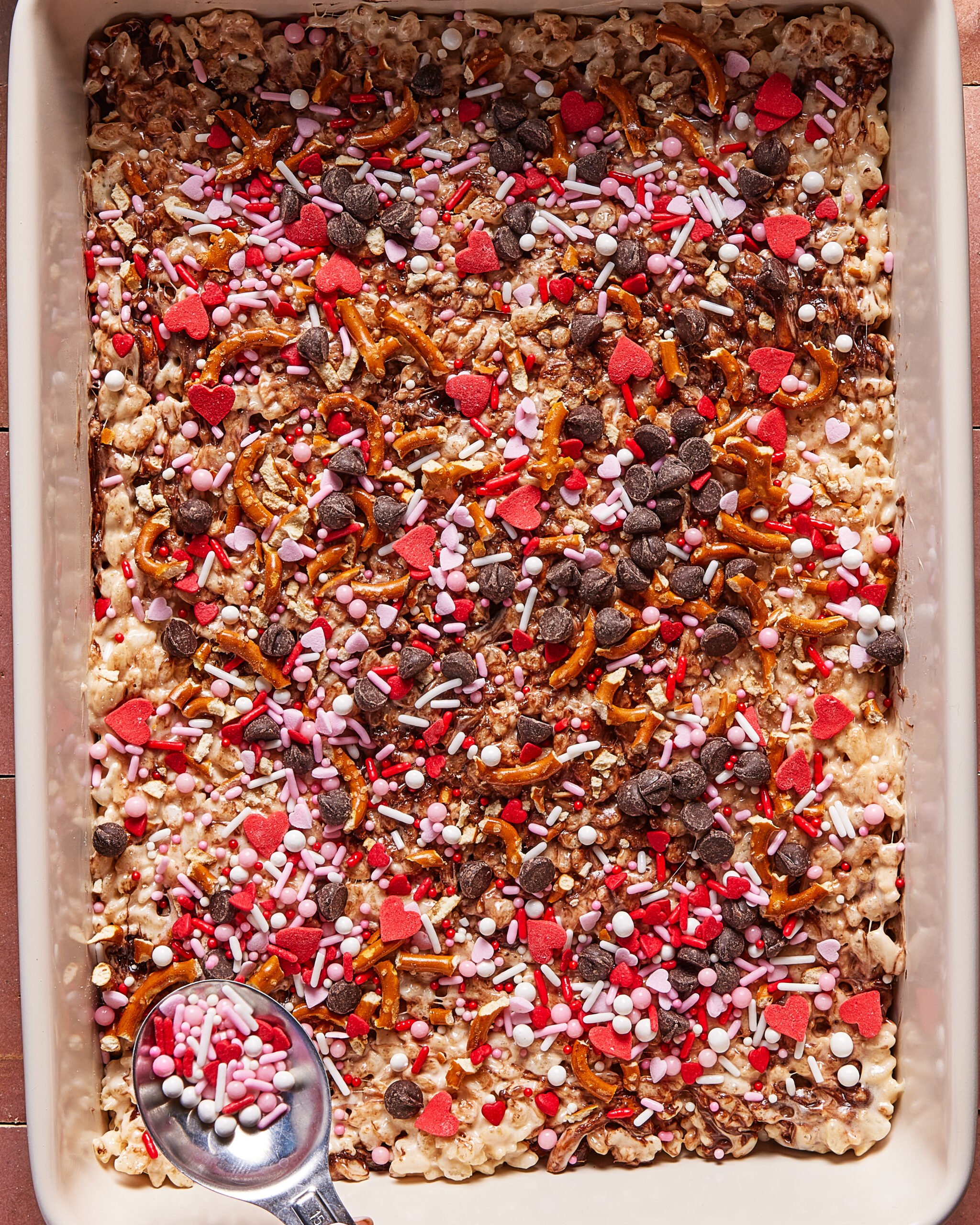 A pan of Rice Krispies treats topped with chocolate chips, pretzel pieces, and colorful heart-shaped and round sprinkles. A spoon in the corner holds extra sprinkles.
