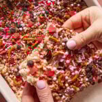 A person holds a decorated dessert bar above a tray of similar bars. The bars are topped with pretzel pieces, heart-shaped sprinkles, colorful round sprinkles, and chocolate chips.