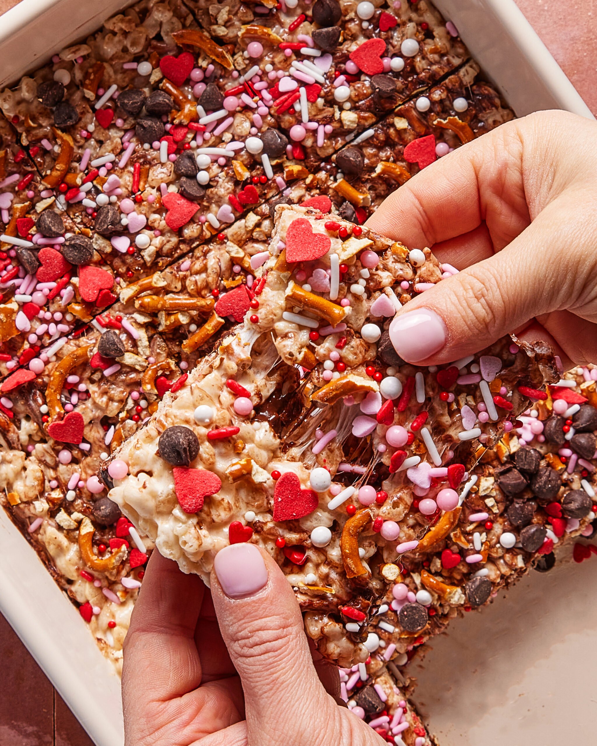 A person holds a decorated dessert bar above a tray of similar bars. The bars are topped with pretzel pieces, heart-shaped sprinkles, colorful round sprinkles, and chocolate chips.