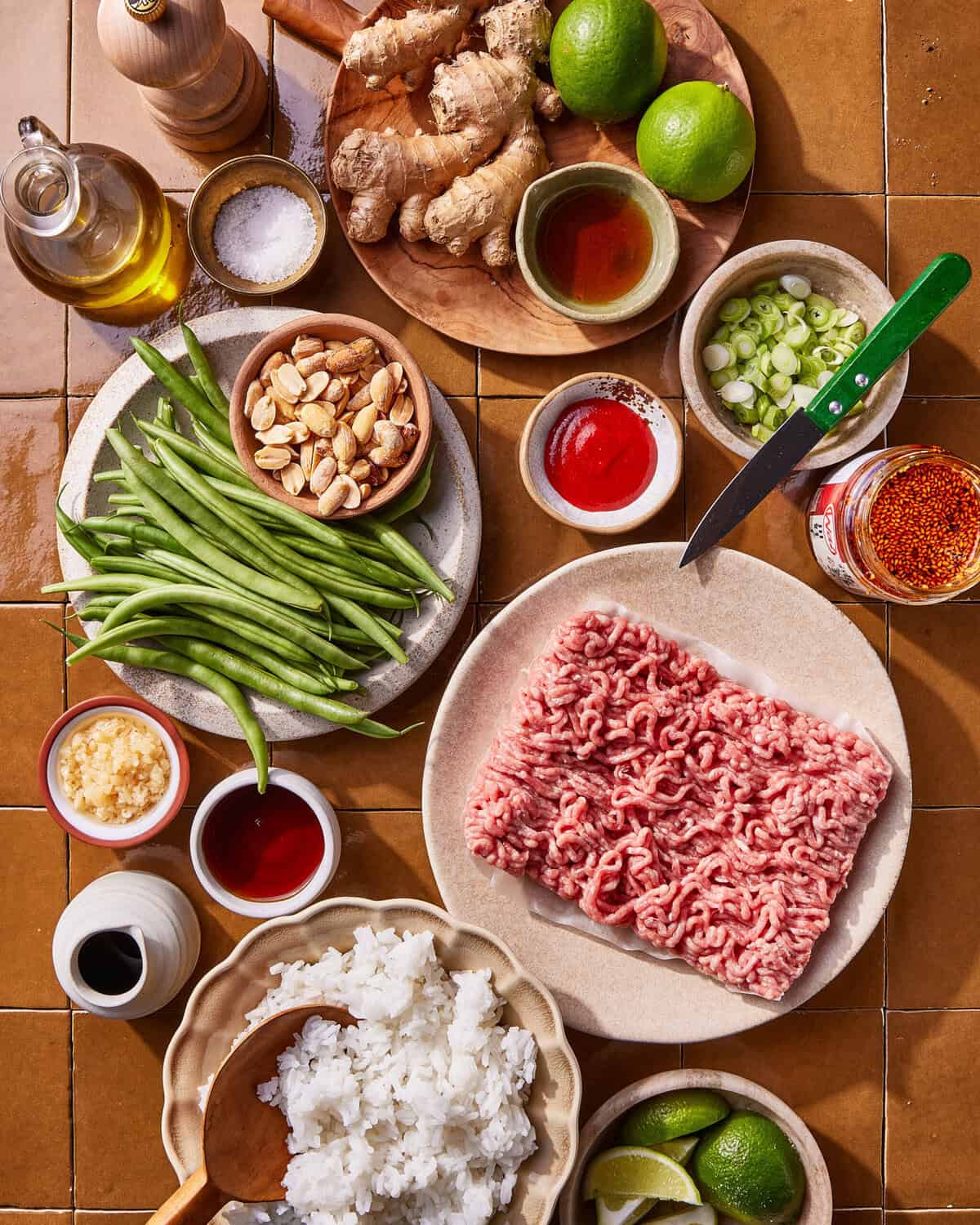 A flat lay of various ingredients on a brown tile surface, including raw ground meat, cooked white rice, green beans, sliced green onions, ginger, limes, almonds, garlic, sauces, oil, and seasonings.