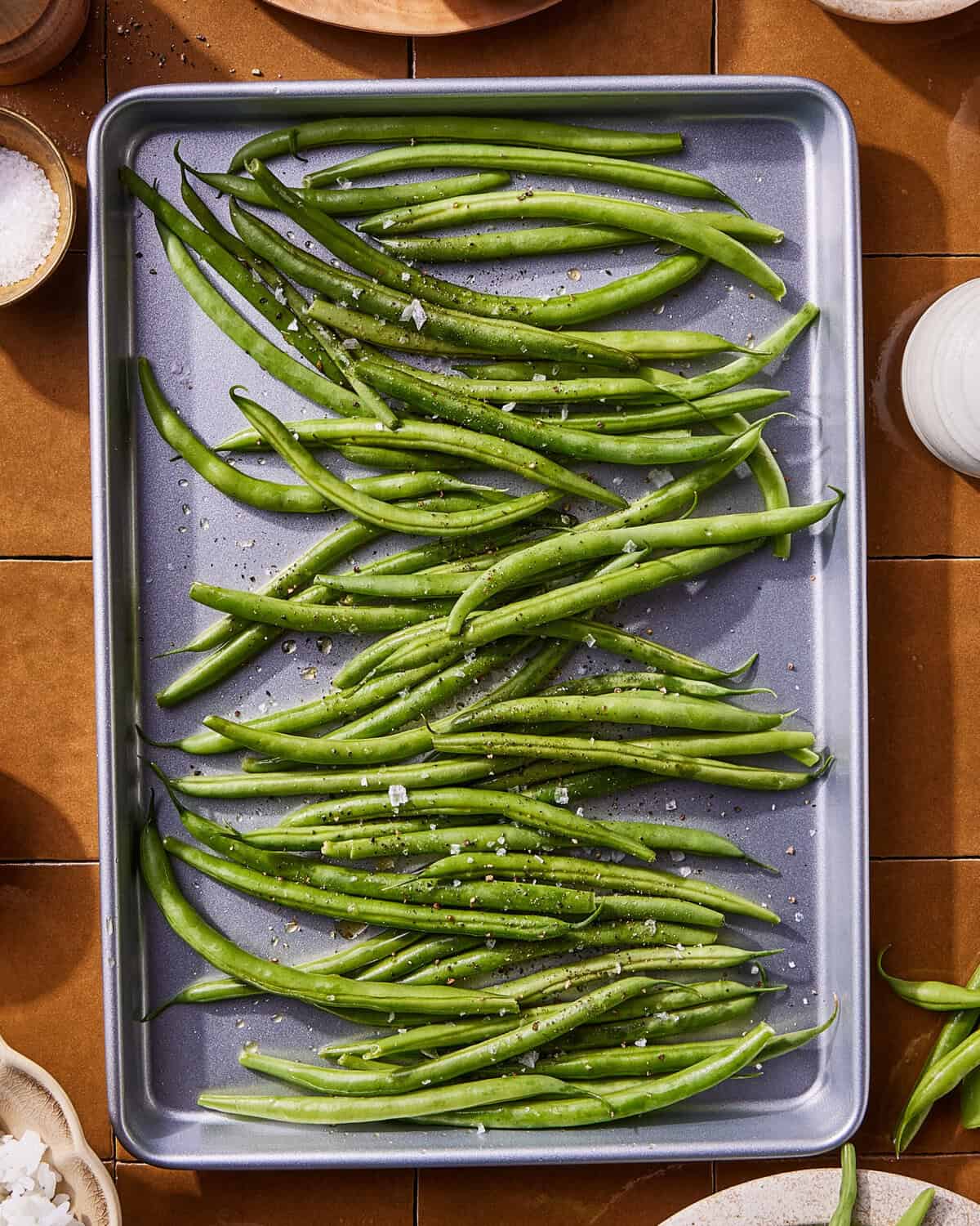 A baking sheet filled with fresh green beans sprinkled with coarse salt and black pepper, ready for roasting. Bowls of salt and other ingredients are visible around the tray on a brown tiled surface.