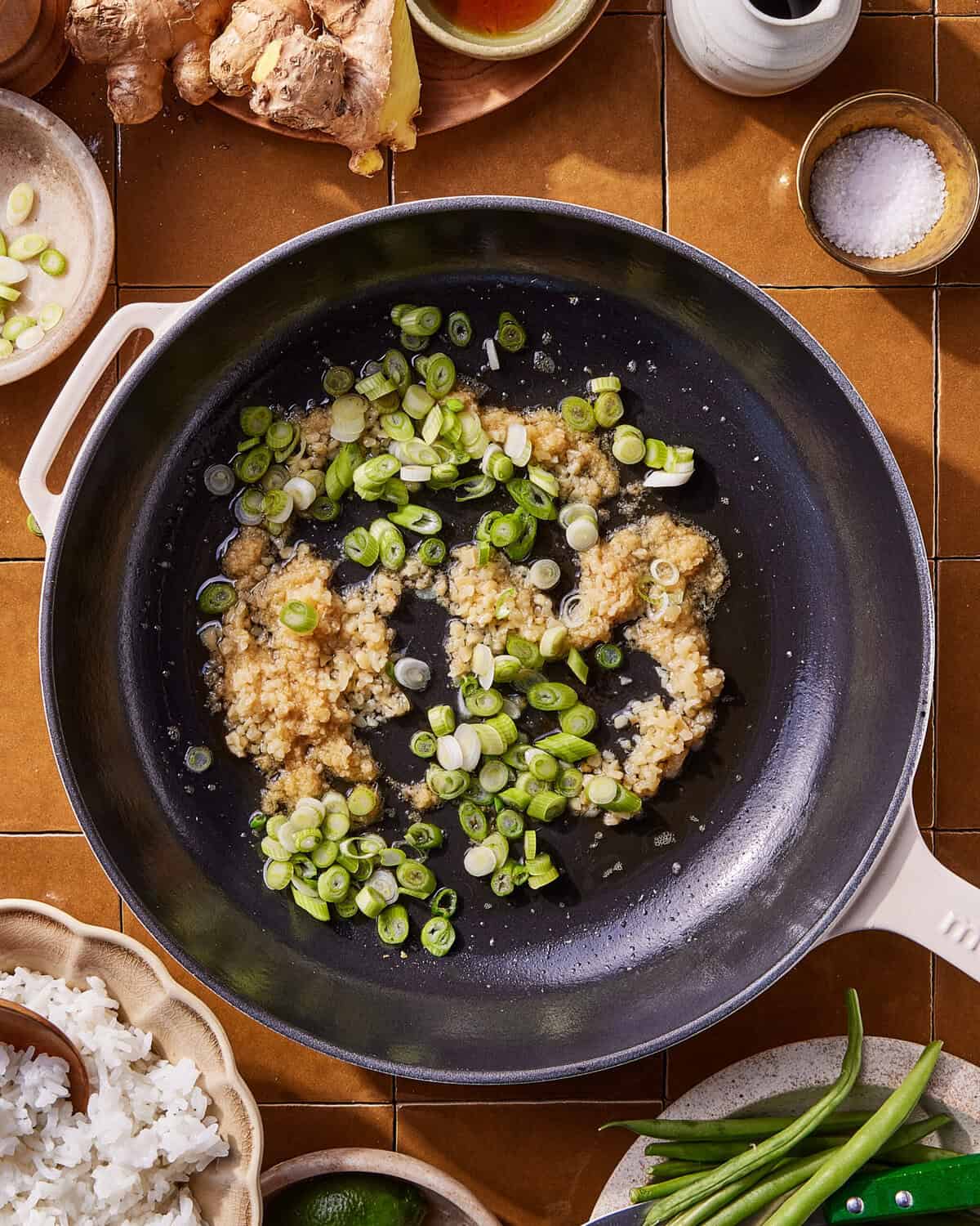 A black skillet with chopped garlic and sliced green onions sizzling in oil, surrounded by bowls of ginger, salt, rice, green beans, and lime on a brown tiled countertop.