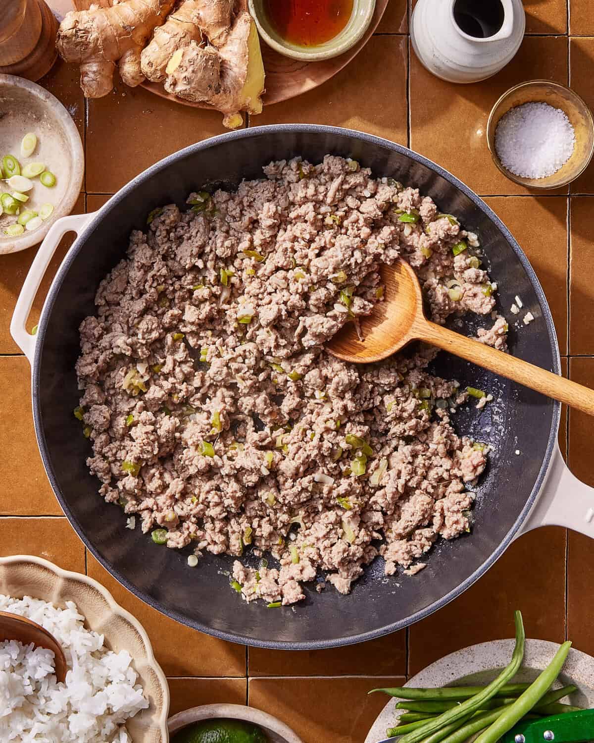 A skillet filled with cooked ground meat mixed with chopped green onions, stirred with a wooden spoon. Surrounding the skillet are ingredients like rice, green beans, ginger, salt, and sauce on a tiled surface.