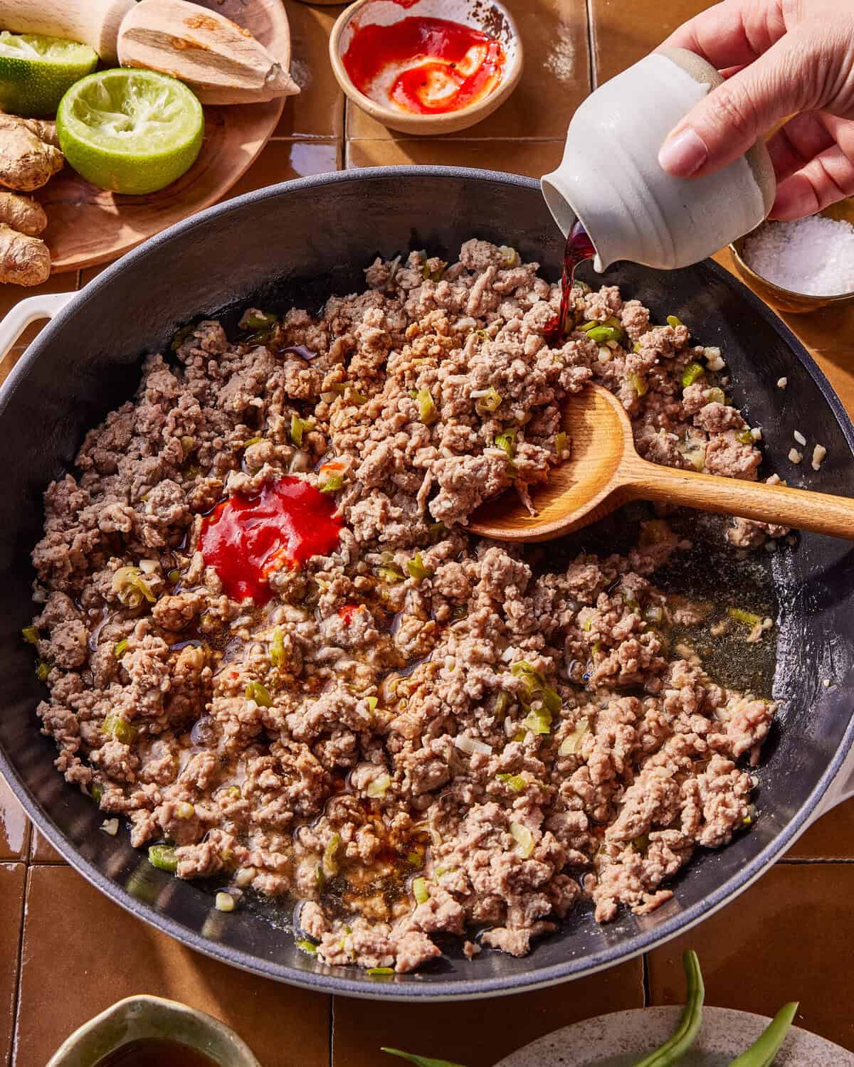 Ground meat cooking in a skillet with chopped vegetables, a wooden spoon, and tomato paste. A hand pours dark liquid from a small pitcher. Ingredients like lime, ginger, and sauce are nearby on a brown tiled surface.