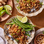 Two bowls of rice topped with ground meat, chopped peanuts, green beans, sliced green onions, and lime wedges. Nearby are a fork, a small dish of peanuts, fresh ginger, and a plate with lime wedges.