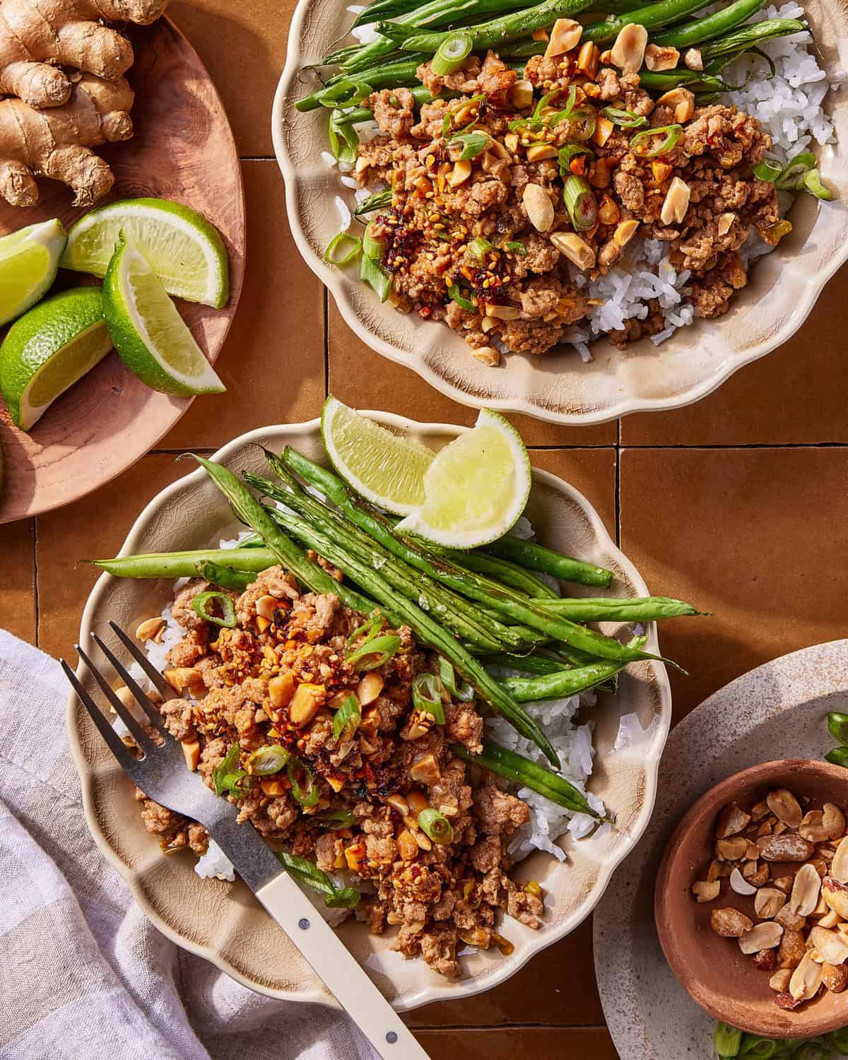 Two bowls of rice topped with ground meat, chopped peanuts, green beans, sliced green onions, and lime wedges. Nearby are a fork, a small dish of peanuts, fresh ginger, and a plate with lime wedges.