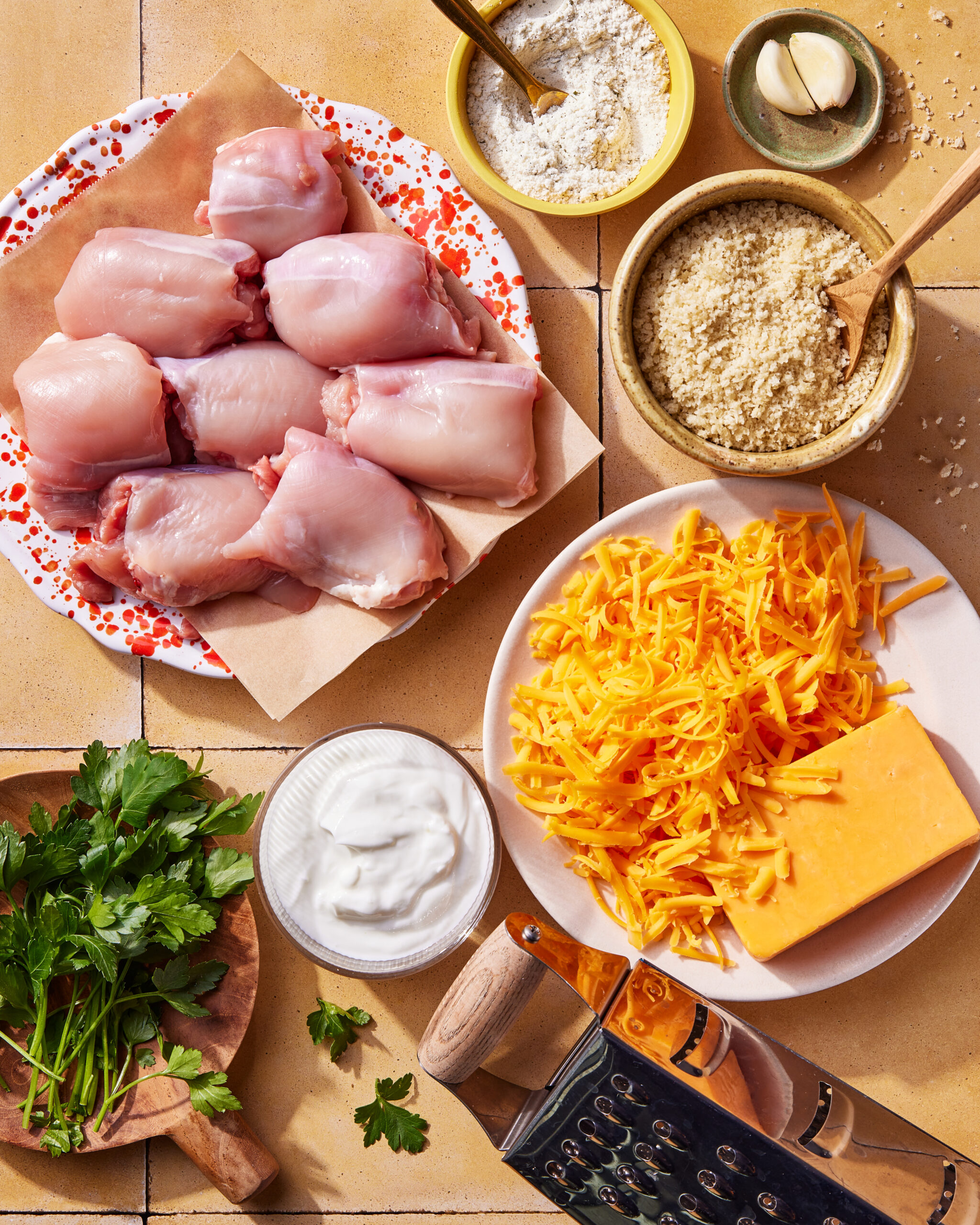 A top view of raw chicken thighs, grated cheddar cheese, a block of cheddar, parsley, sour cream, panko crumbs, flour, a garlic clove, and a grater arranged on a tiled surface.