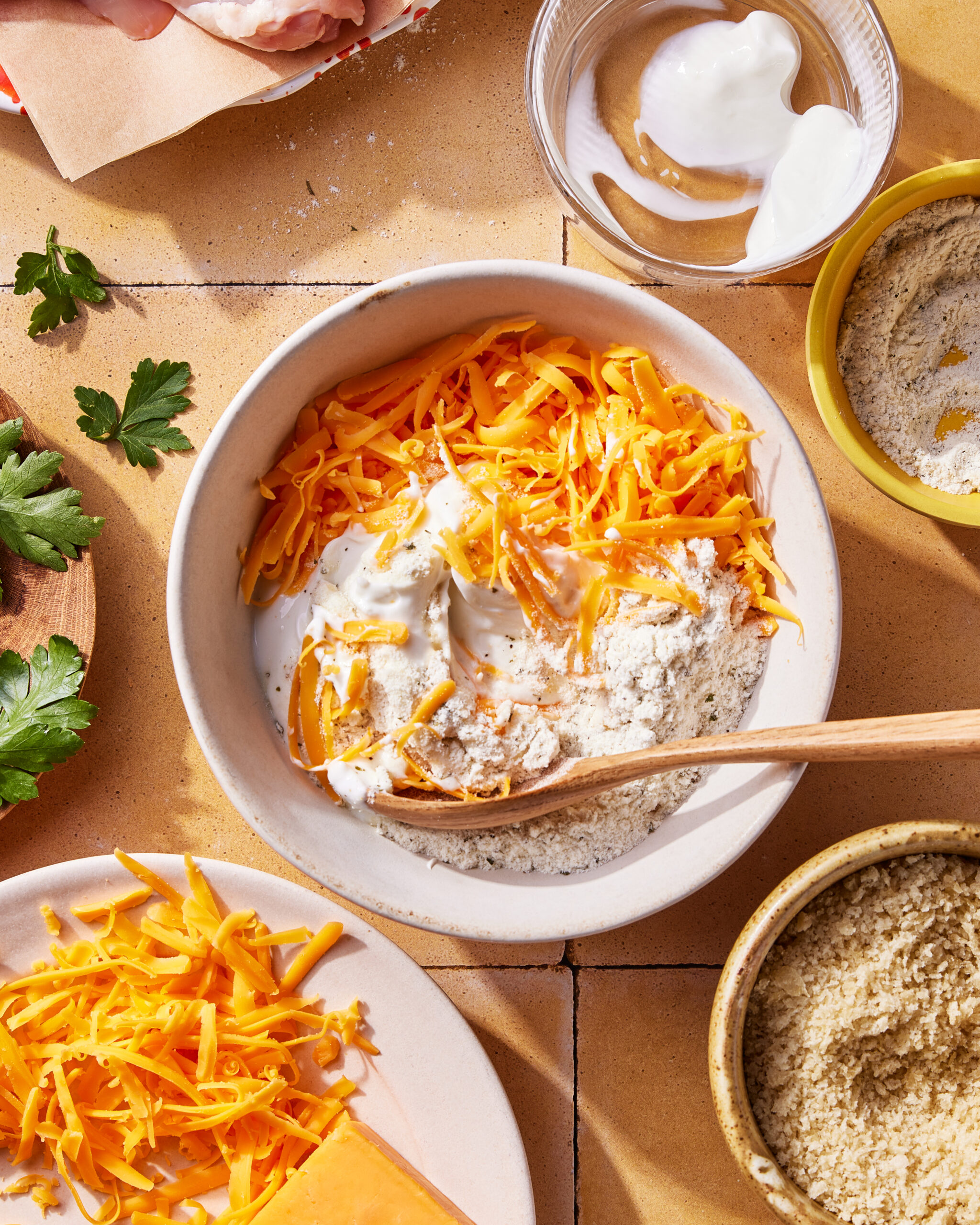A bowl with shredded cheddar cheese, flour, and sour cream being mixed with a wooden spoon. Surrounding the bowl are parsley, more shredded cheese, sour cream, and flour in separate bowls, all on a tile surface.