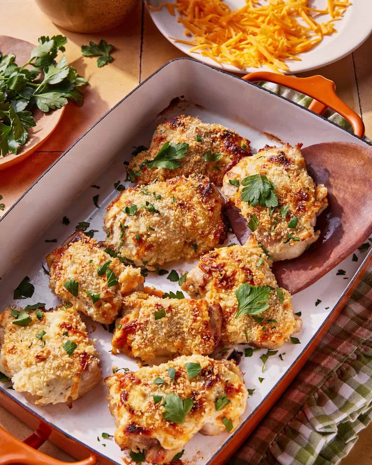 A baking dish with eight pieces of baked chicken topped with golden breadcrumbs and melted cheese, garnished with fresh parsley. Plates of shredded cheese and fresh herbs are visible in the background.