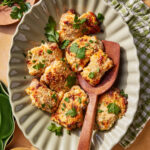 A platter of baked chicken pieces topped with golden, melted cheese and garnished with fresh parsley, served on a wavy-edge plate with a wooden serving spoon. A green checkered towel and herbs are nearby.