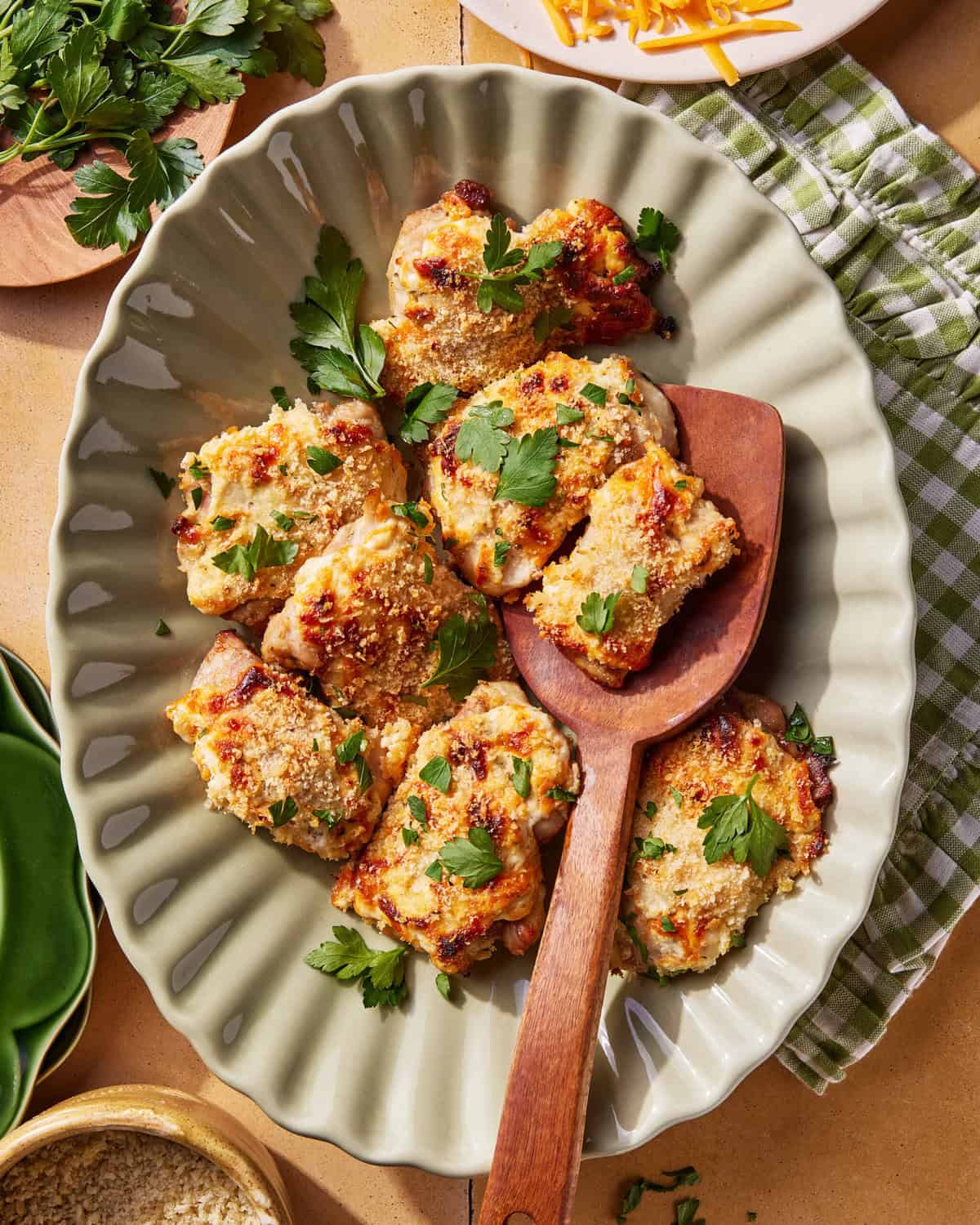 A platter of baked chicken pieces topped with golden, melted cheese and garnished with fresh parsley, served on a wavy-edge plate with a wooden serving spoon. A green checkered towel and herbs are nearby.