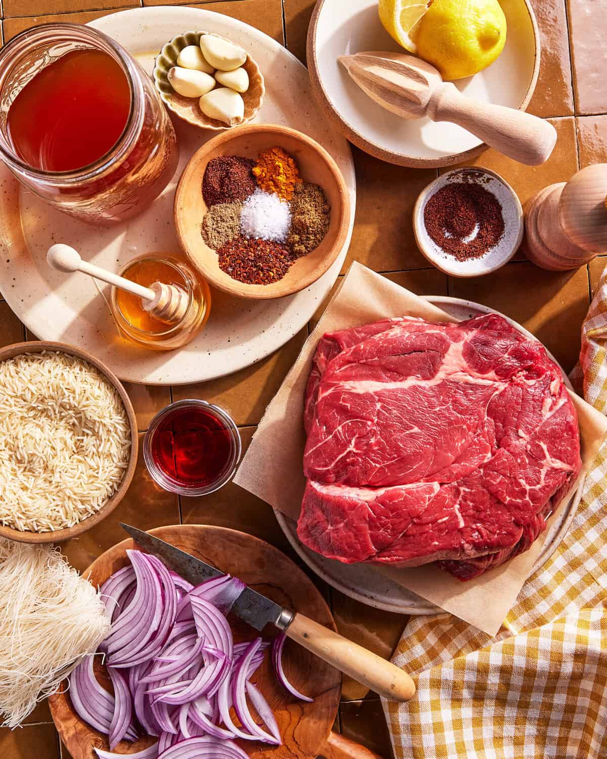 A top view of raw beef, sliced red onions, rice, spices, garlic, honey, lemon, broth, and sumac arranged on a table, ready for cooking. A checkered napkin and a wooden-handled knife are also visible.