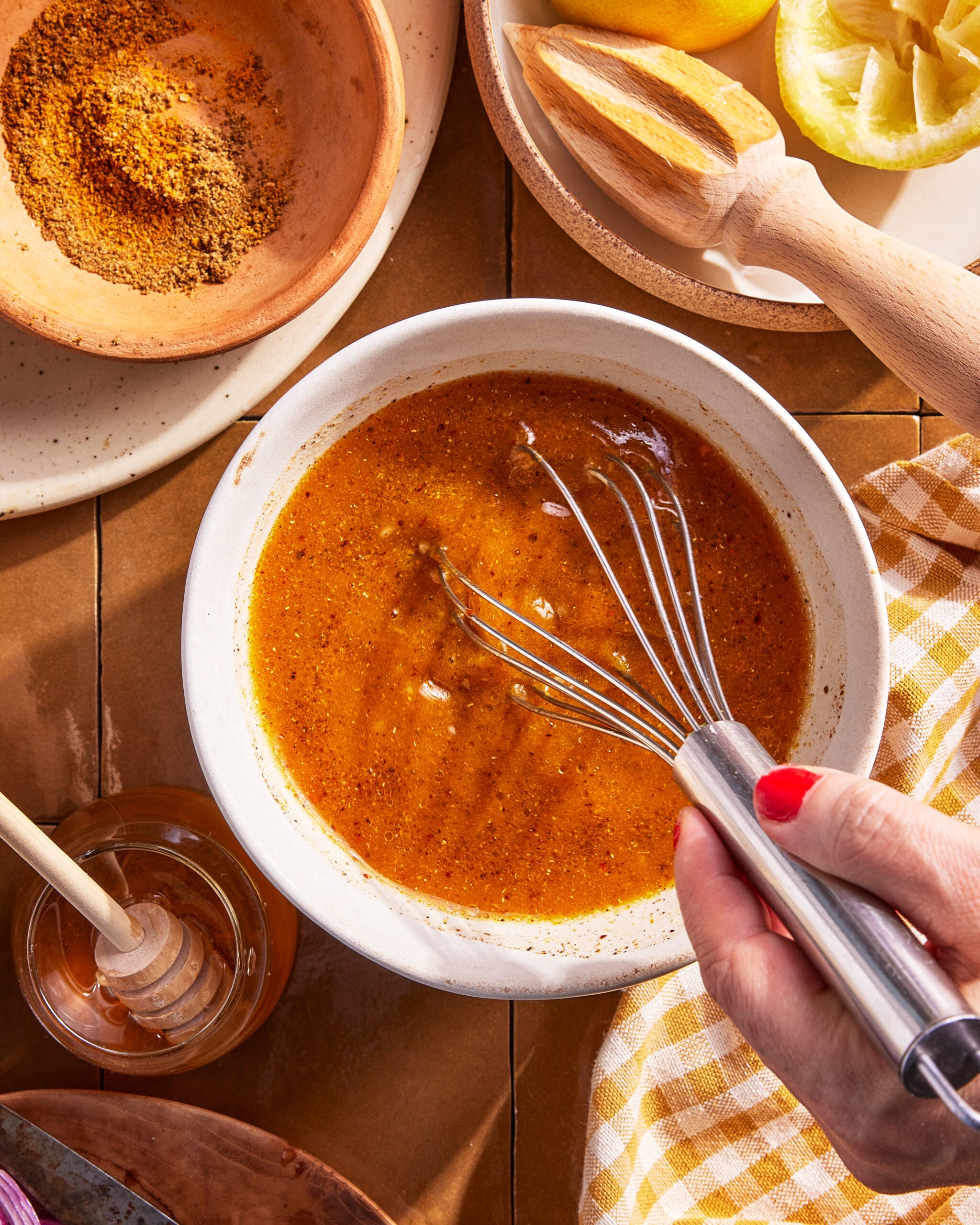 A hand with red nail polish uses a whisk to mix a golden-brown dressing in a white bowl, surrounded by honey, spices, lemons, and a checkered cloth on a wooden surface.
