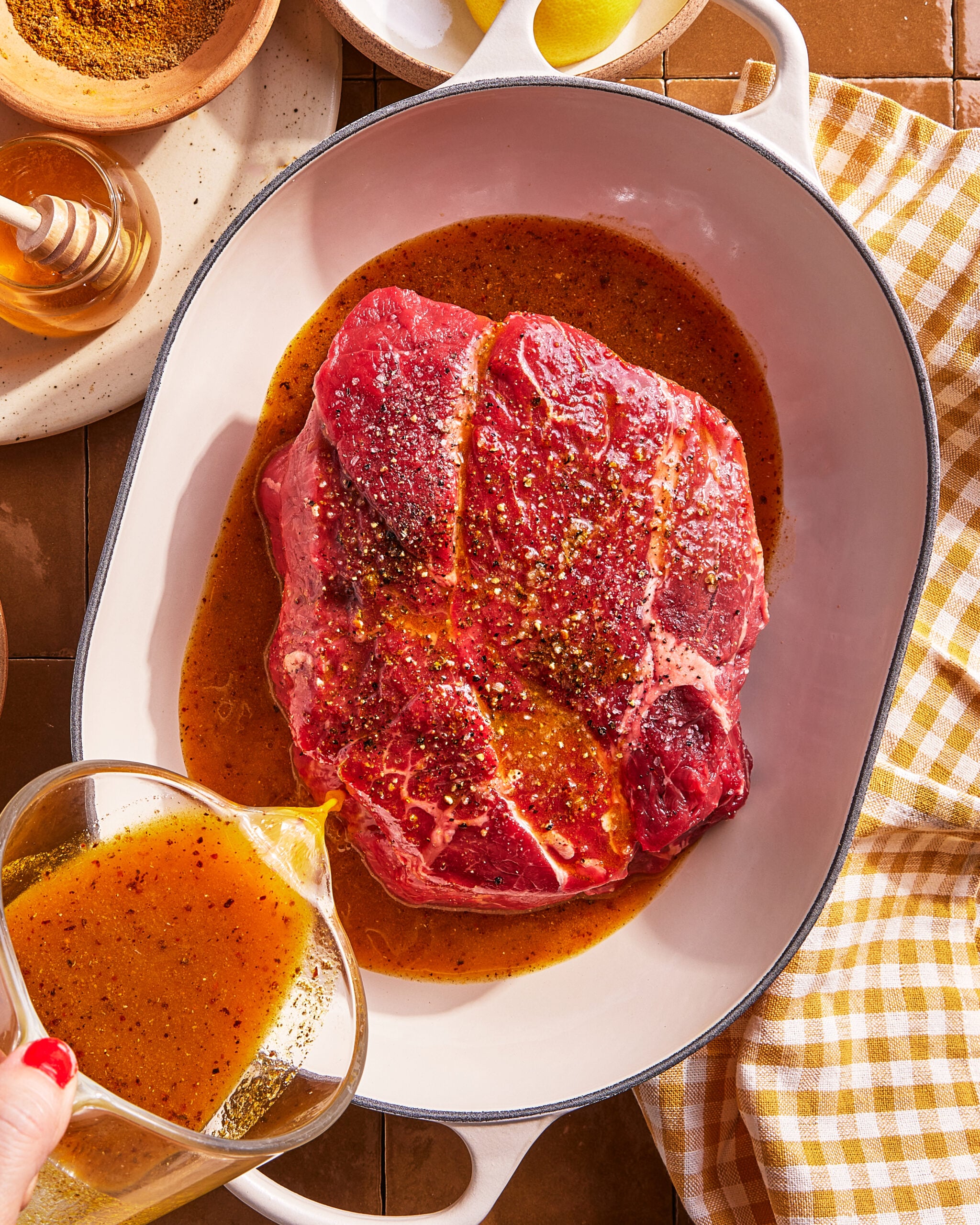 A raw seasoned beef roast sits in a white oval dish as someone pours a golden marinade over it. Nearby are a jar of honey, a bowl of spices, lemon wedges, and a yellow gingham cloth.