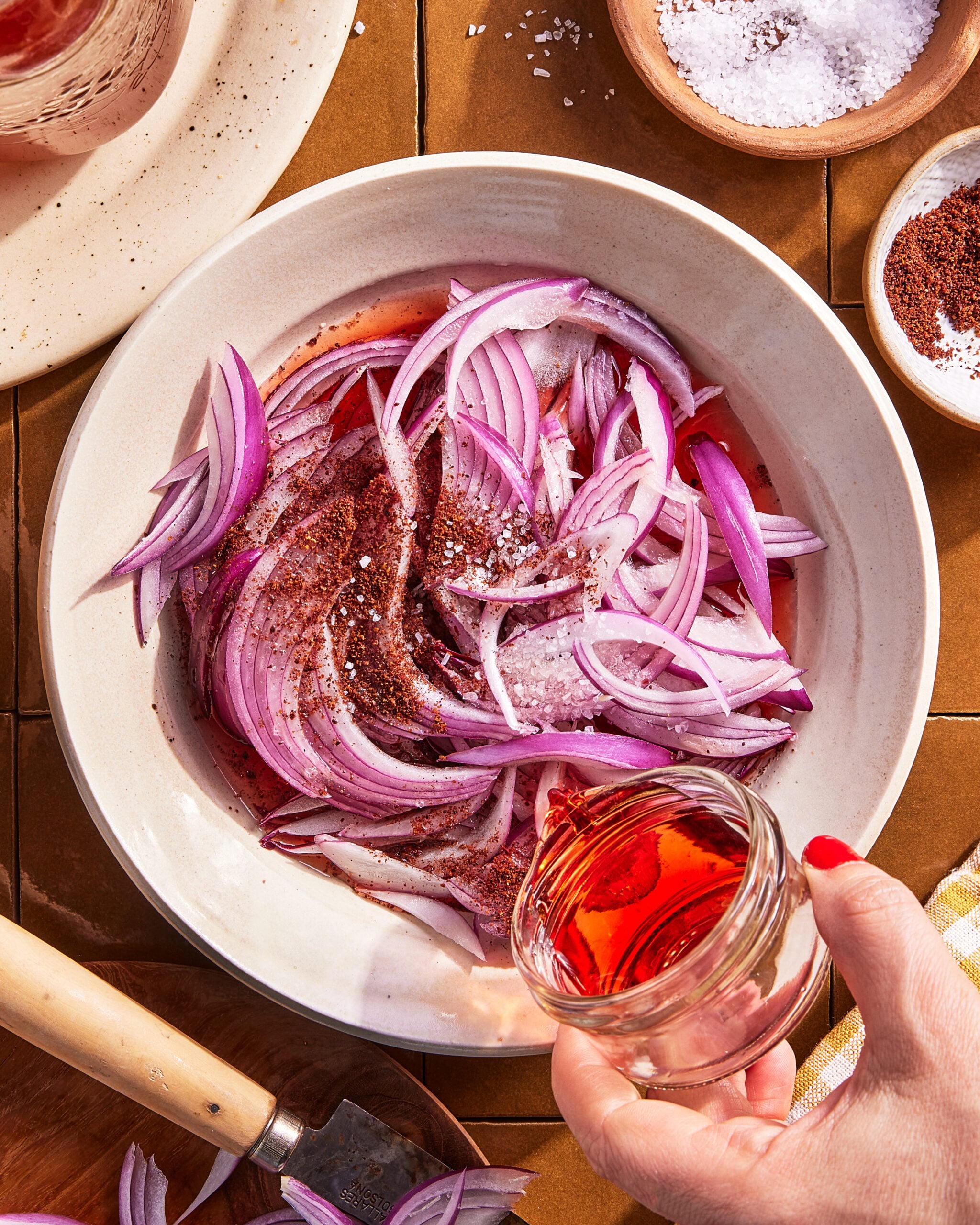 A hand pours red liquid, likely vinegar, into a bowl of thinly sliced red onions sprinkled with salt and spices. Small bowls of salt and a spice are nearby on a brown tiled surface.