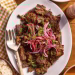A white plate with shredded, seasoned beef topped with sliced red onions and fresh herbs, next to a fork. A bowl of rice and a small dish with herbs are nearby on a brown table with a checkered napkin.