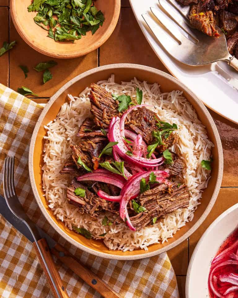 A bowl of white rice topped with shredded beef, pickled red onions, and fresh herbs. A fork and knife are nearby, along with a bowl of chopped herbs and a plate of cooked meat.
