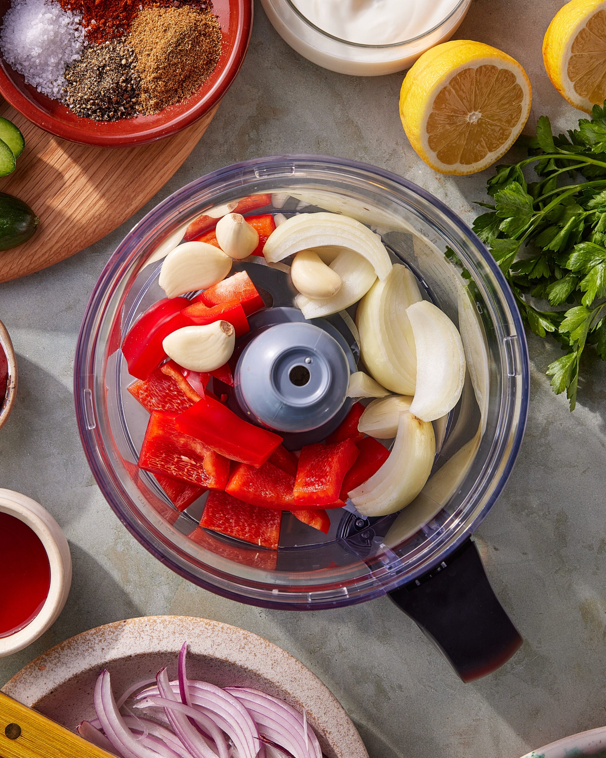 A food processor bowl filled with garlic cloves, red bell pepper pieces, and onion wedges, surrounded by fresh lemon, parsley, spices, red onion slices, sour cream, and other ingredients on a countertop.