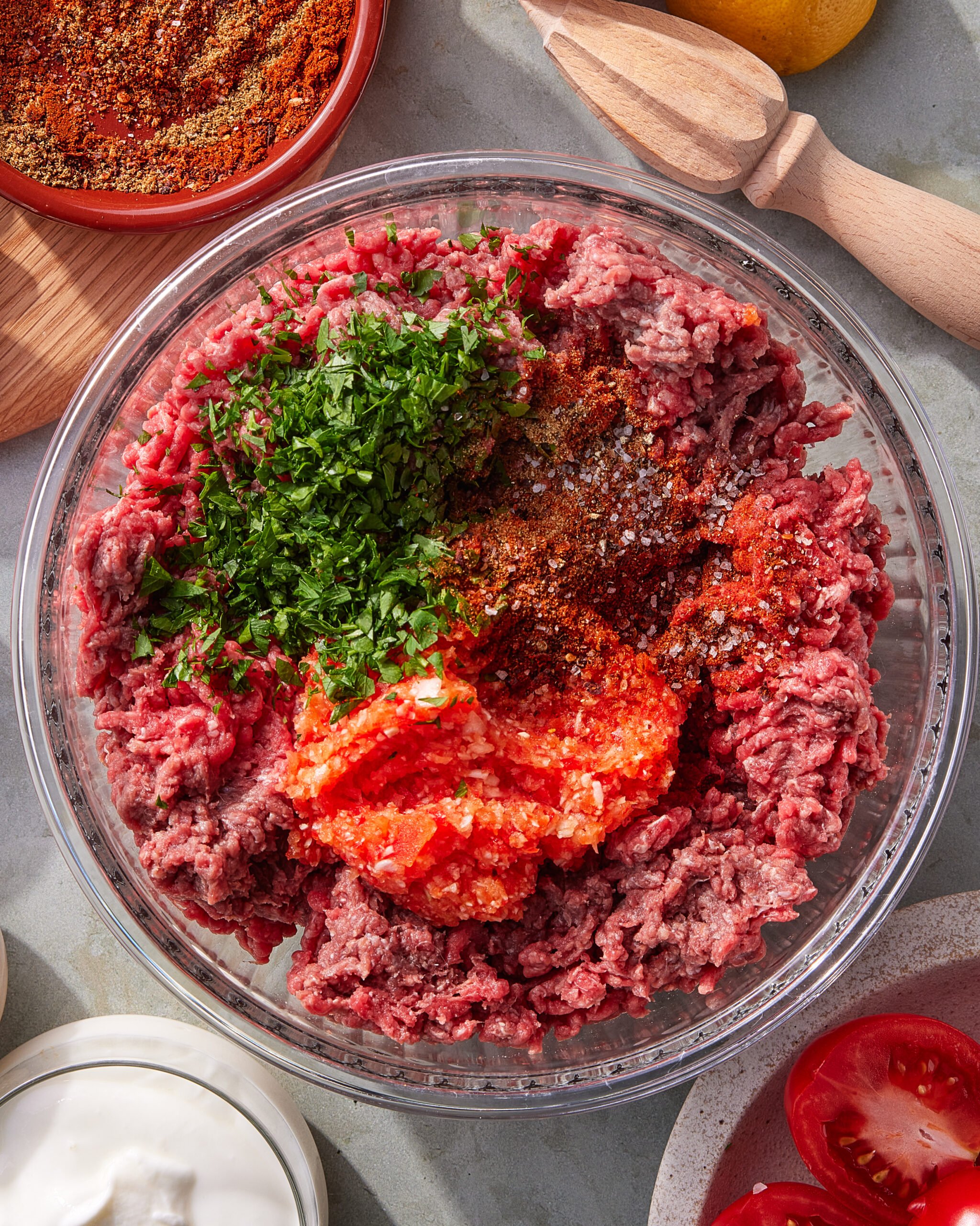 A glass bowl filled with raw ground beef, topped with chopped herbs, red spices, and a dollop of red sauce, sits on a table surrounded by ingredients like tomatoes, sour cream, and a citrus reamer.