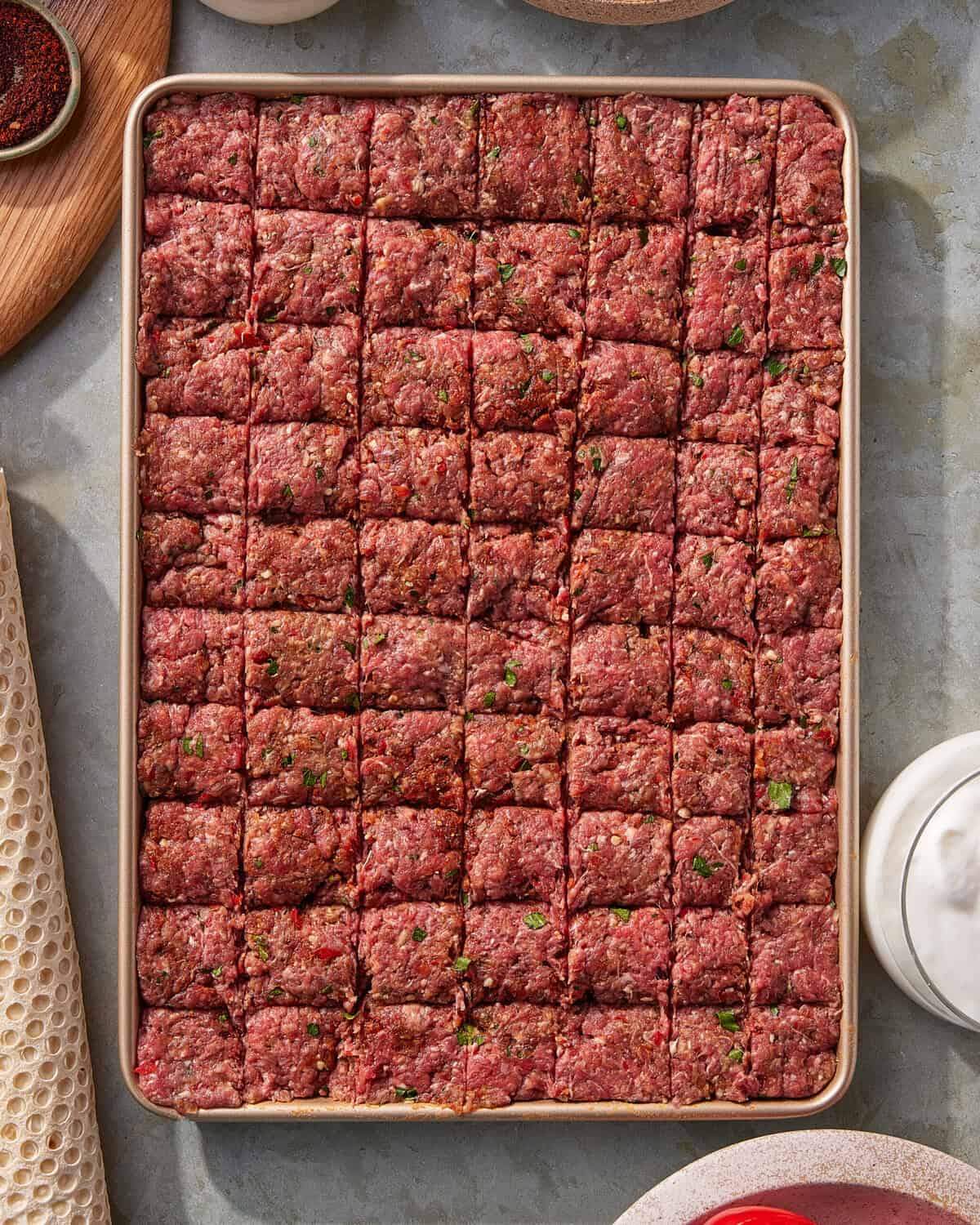 A baking sheet filled with evenly cut squares of seasoned ground meat, arranged in a grid pattern. The meat mixture appears to include green herbs and spices. Various kitchen items are visible around the edges.