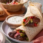 Two rolled flatbreads filled with grilled meat, chopped tomatoes, cucumbers, and red onions are served on a plate with a side of white dipping sauce, with extra salad and flatbread in the background.