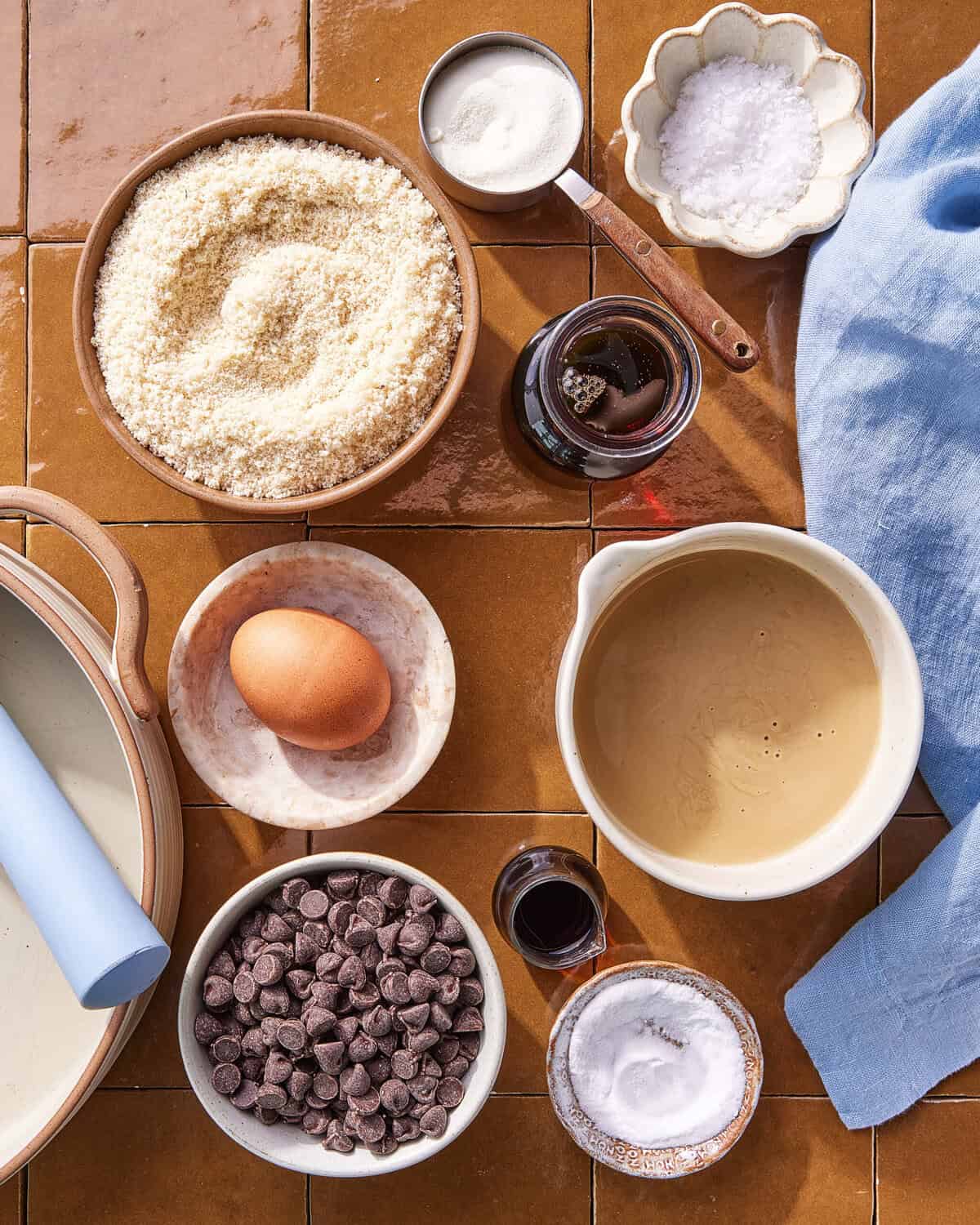 A top-down view of baking ingredients on a brown tile surface, including almond flour, an egg, chocolate chips, honey, salt, vanilla extract, baking soda, and a blue cloth napkin.