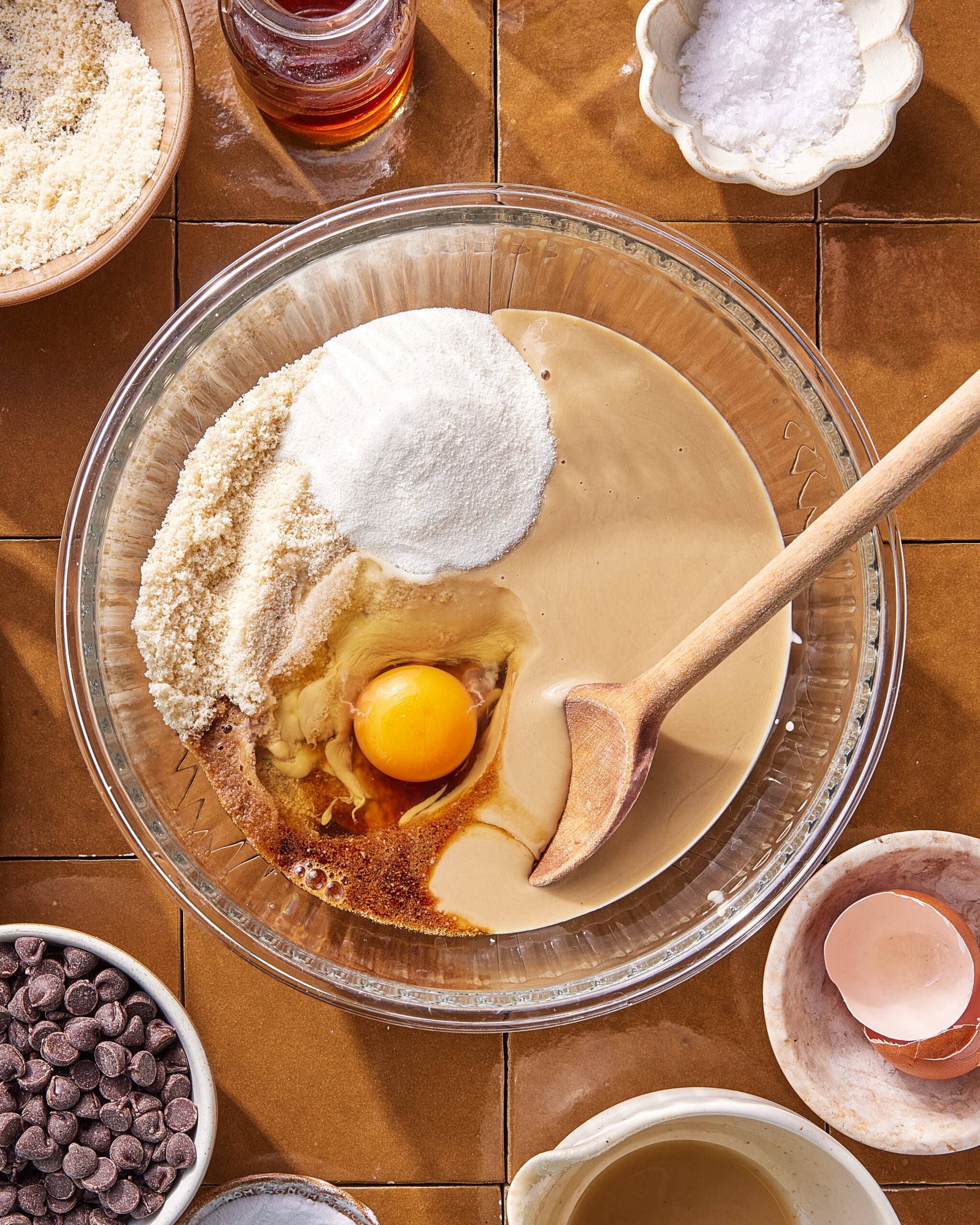 A glass bowl with tahini, sugar, almond flour, an egg, and vanilla, ready to be mixed with a wooden spoon. Surrounding the bowl are chocolate chips, salt, almond flour, and an empty eggshell.