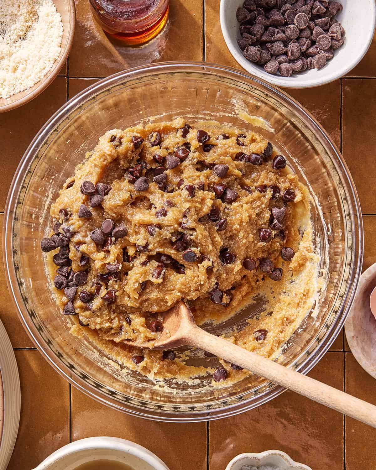 A glass bowl filled with chocolate chip cookie dough and a wooden spoon, surrounded by ingredients like chocolate chips, an egg, almond flour, and a bottle of vanilla on a brown tiled surface.